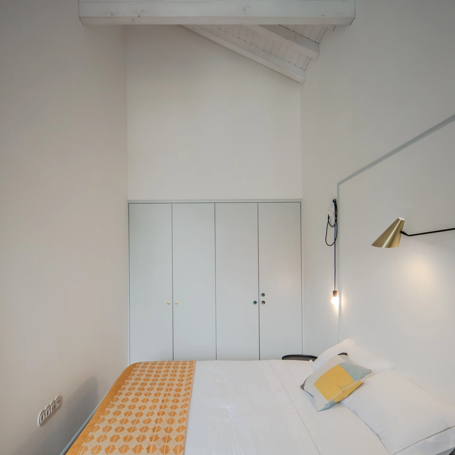 A minimalist white bedroom featuring a bed with an orange geometric throw, built-in wardrobes, and a vaulted ceiling with exposed white beams.