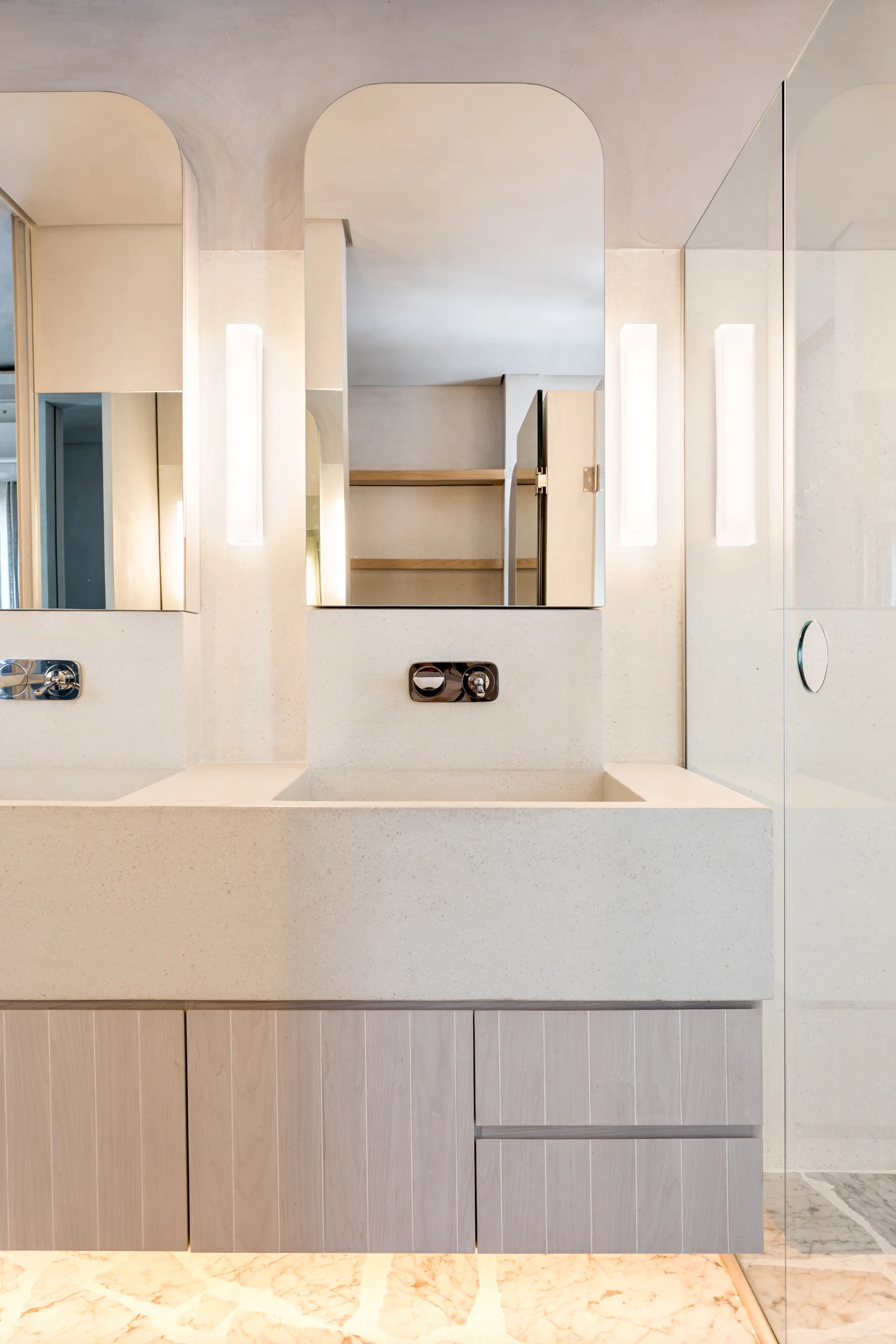 A modern bathroom double vanity features integrated stone sinks, vertical ribbed wood cabinets, arched mirrors, and glowing wall sconces.