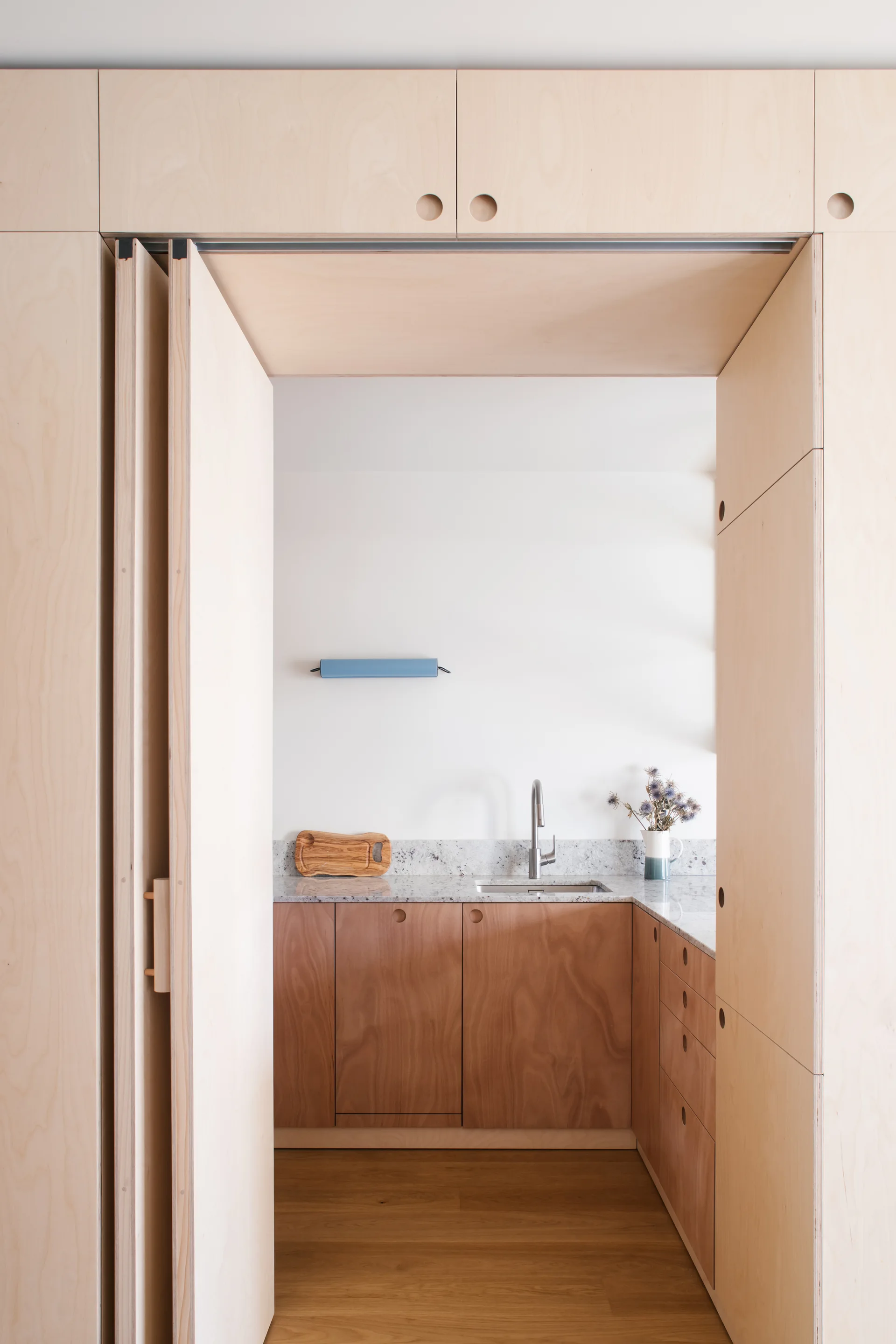 A view through a plywood-paneled doorway reveals a minimalist kitchen with warm wood lower cabinets and a speckled stone countertop.