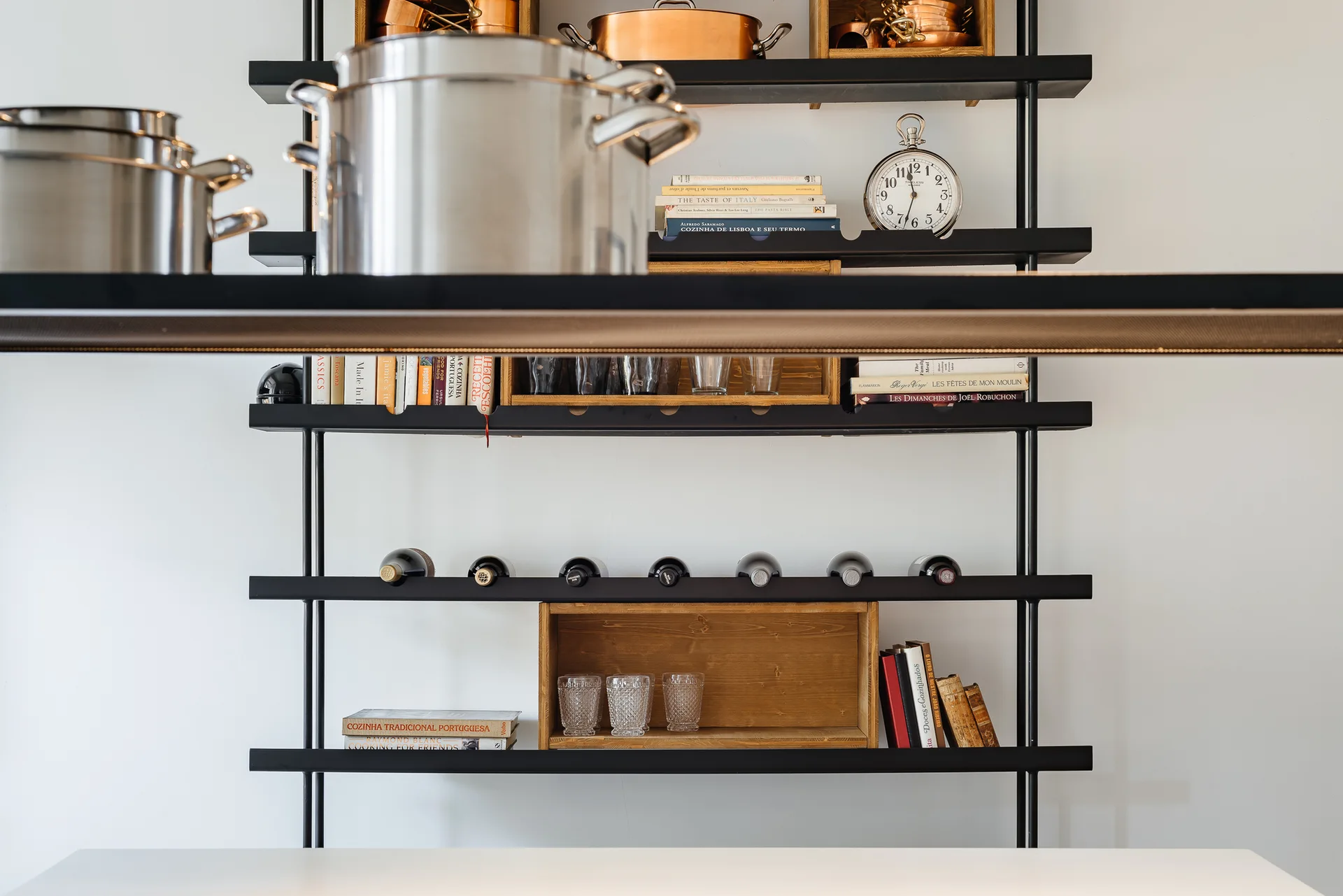 A black metal open shelving unit mounted on a white wall holds stainless steel and copper pots, wine bottles, books, and wooden crates.