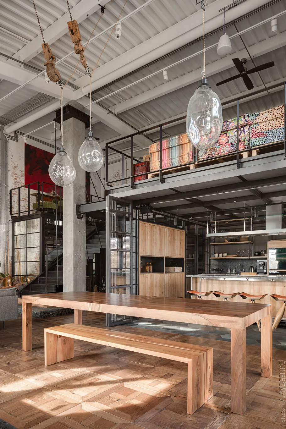 An industrial loft dining space features a long wooden table and bench, oversized glass pendant lights, and an overhead mezzanine level.