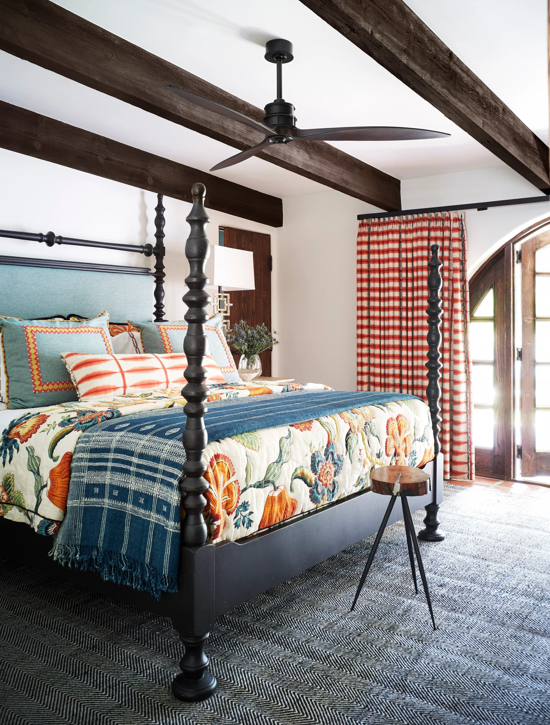 An eclectic bedroom featuring a black spindle four-poster bed with colorful bedding beneath dark wood ceiling beams and near red plaid curtains.