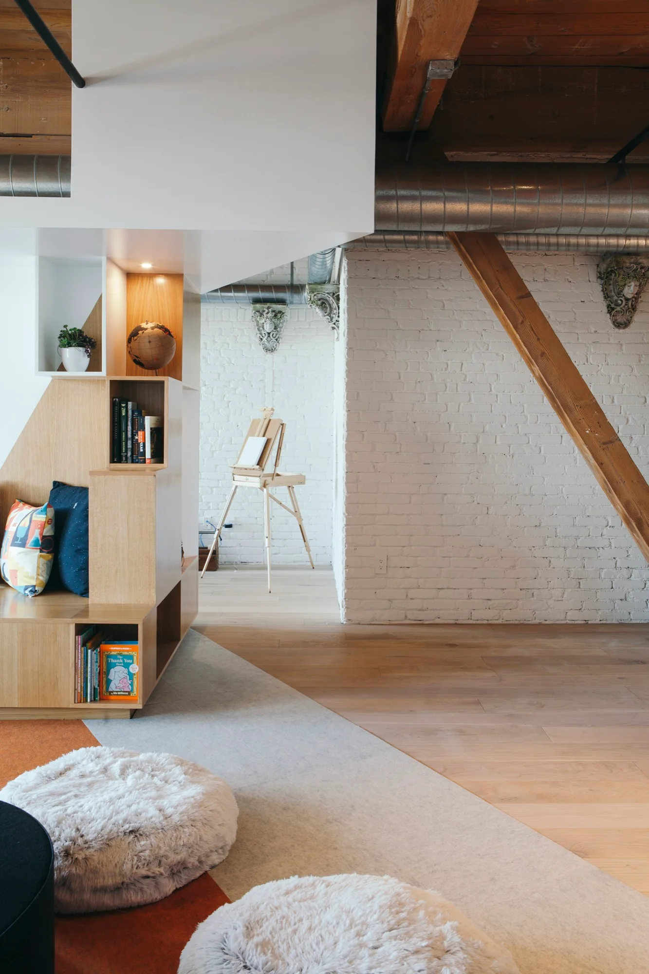 An industrial loft interior features a custom wooden shelving unit with a bench, a white brick wall, exposed timber beams, and an artist easel.