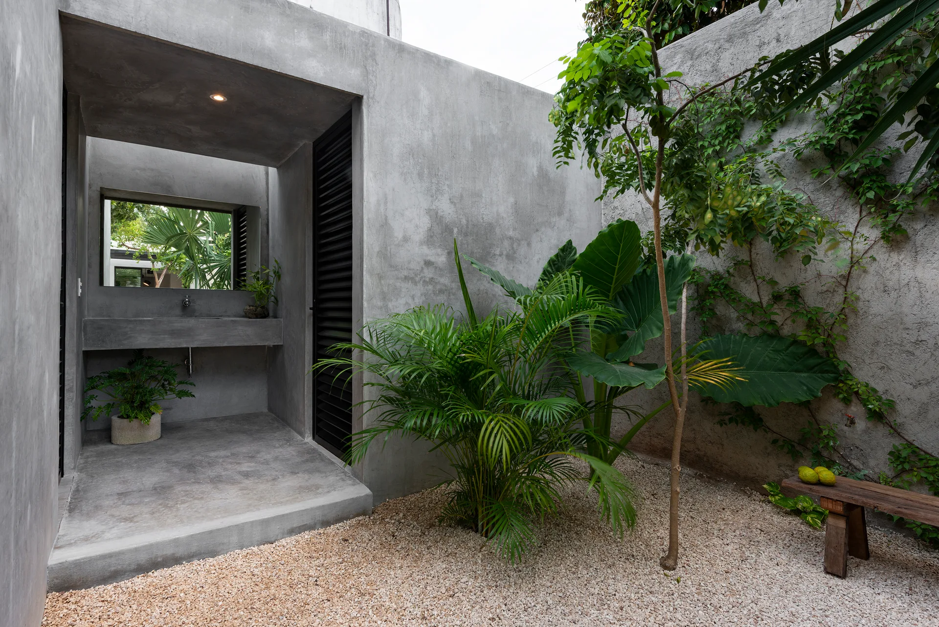 A grey concrete outdoor sink alcove with a mirror overlooks a gravel courtyard filled with leafy green tropical plants and a small wooden bench.