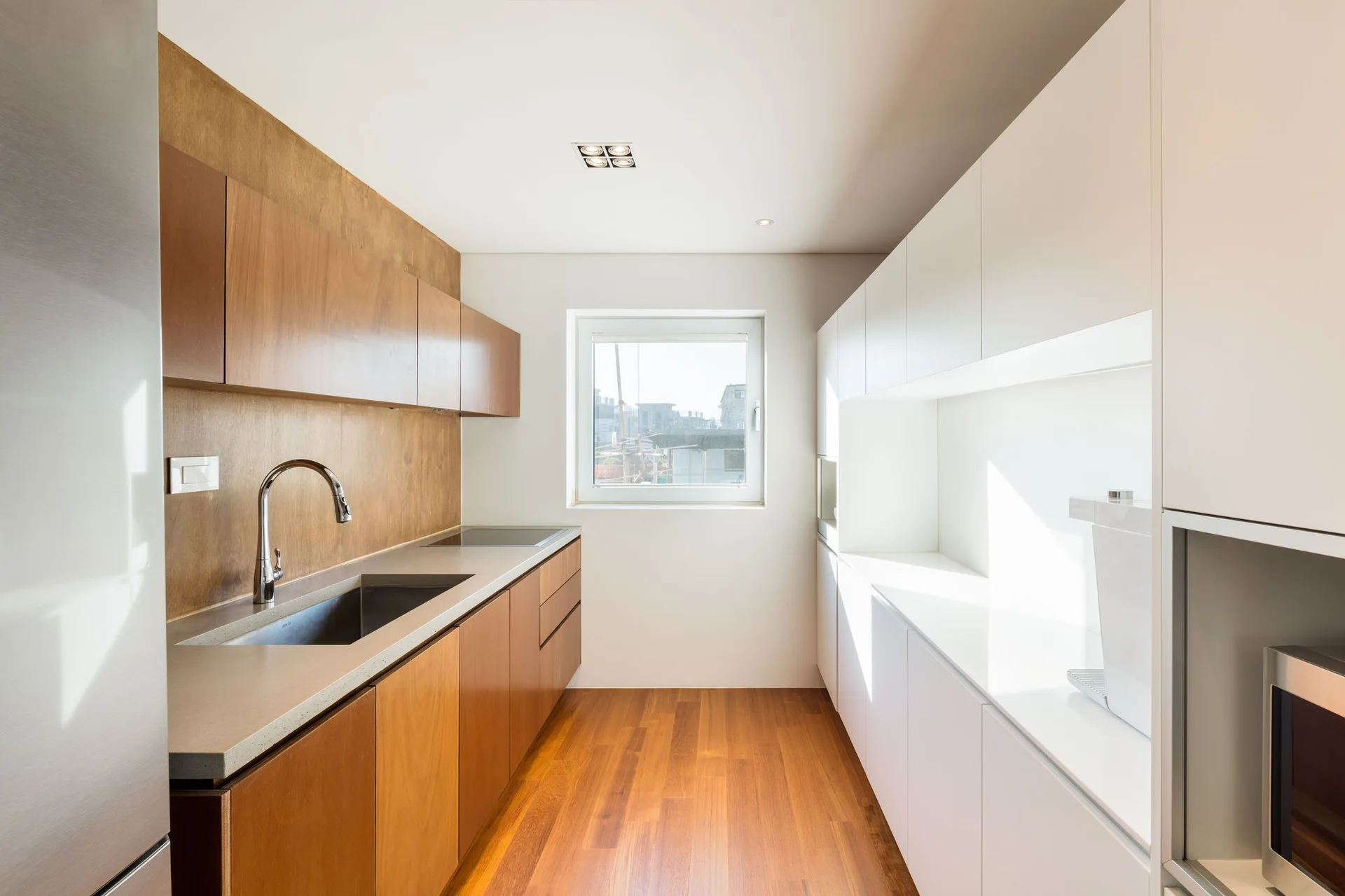 A minimalist galley kitchen with warm wood cabinets on one side, seamless white cabinets on the other, a stainless steel sink, and wood flooring.