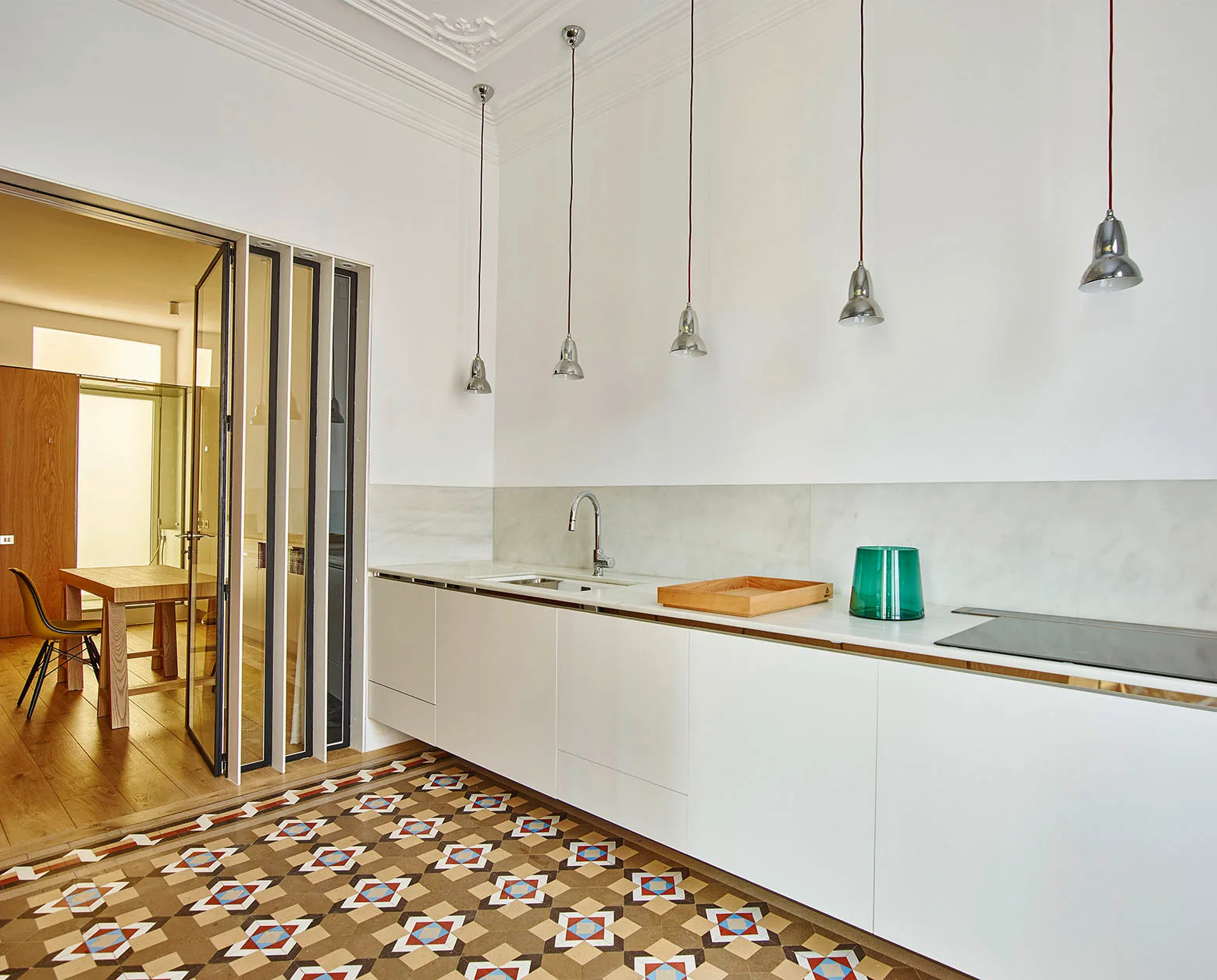 A modern white kitchen featuring flat-front cabinets, chrome pendant lights, and decorative geometric floor tiles leading to a dining room.