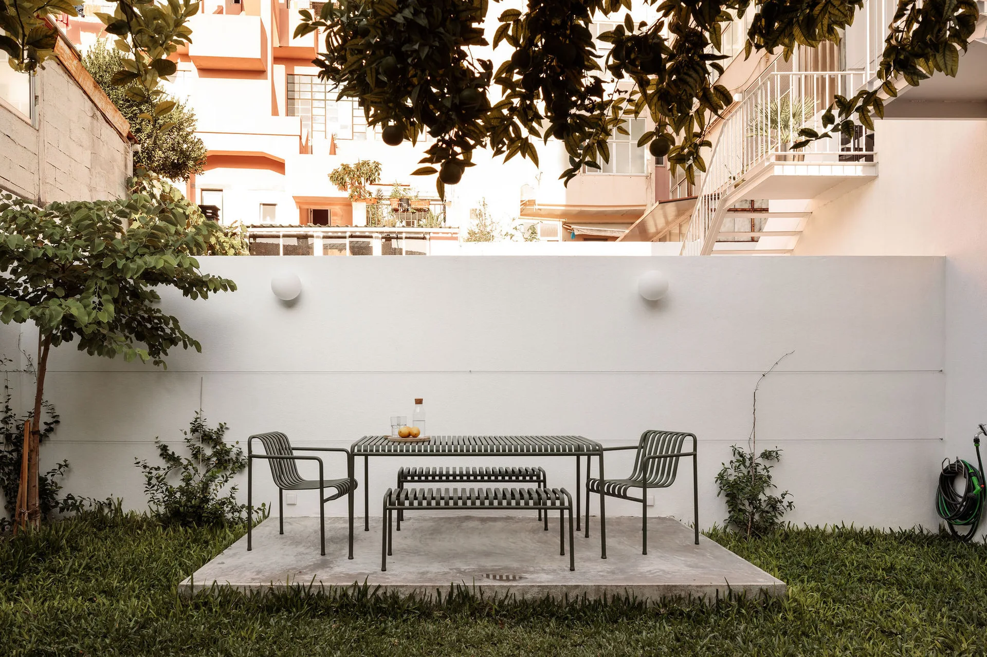 Sage green slatted metal dining furniture sits on a rectangular concrete slab in a grassy backyard against a white wall.