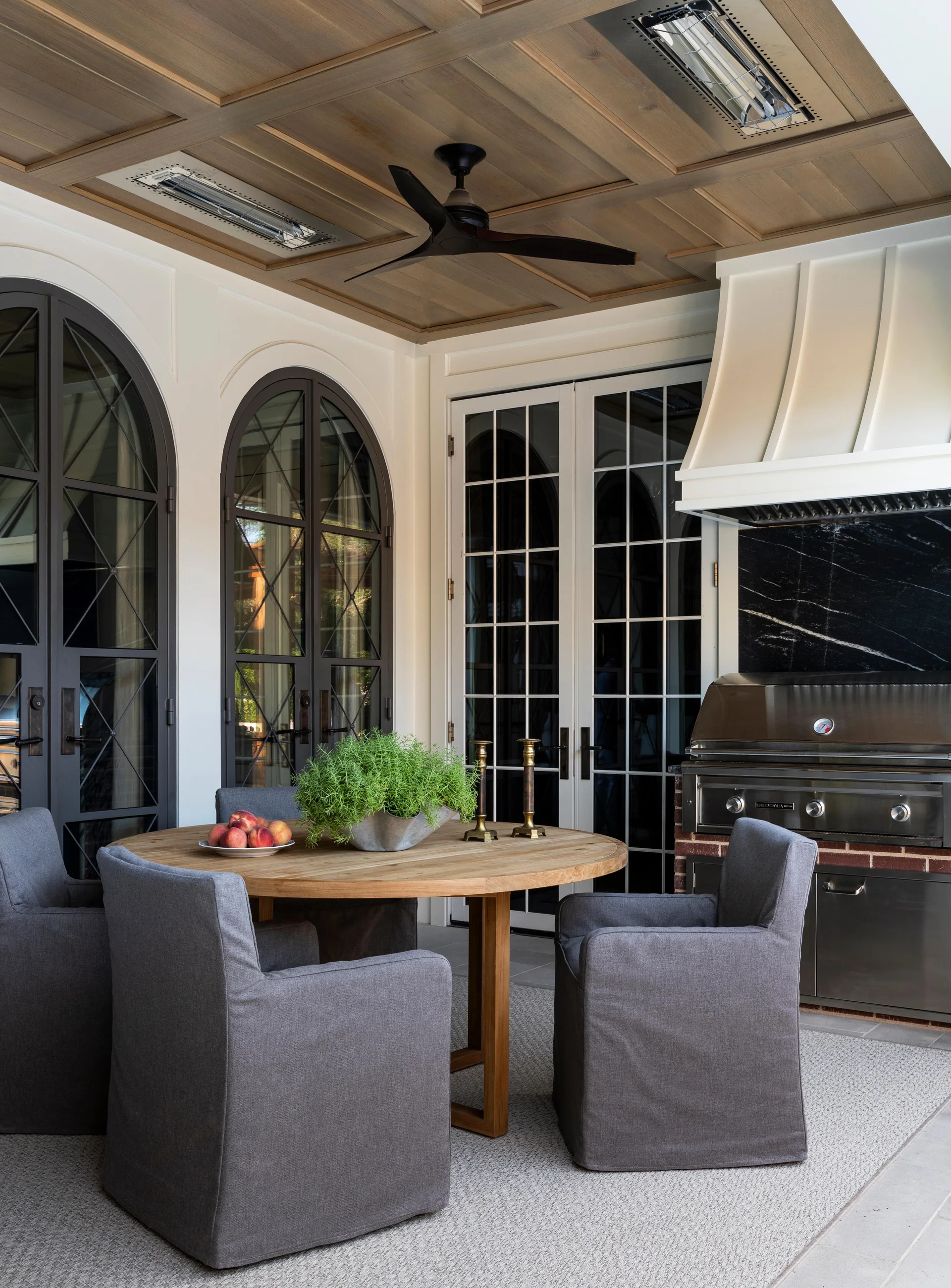 A covered patio with a round wood dining table, gray slipcovered chairs, and an outdoor kitchen under a paneled wood ceiling.