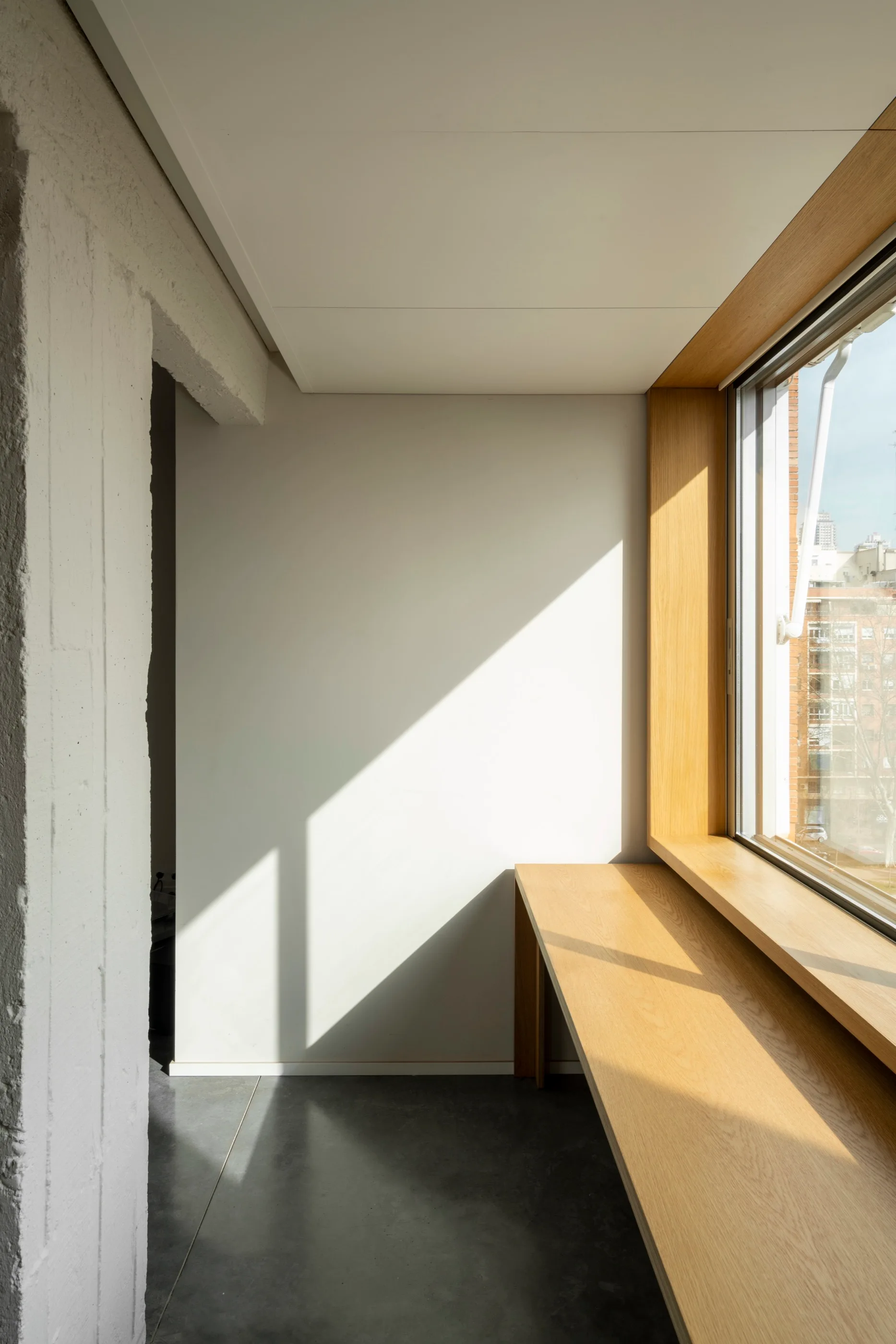 A custom wooden desk is integrated with a deep windowsill next to a painted concrete column and white walls with cast shadows.