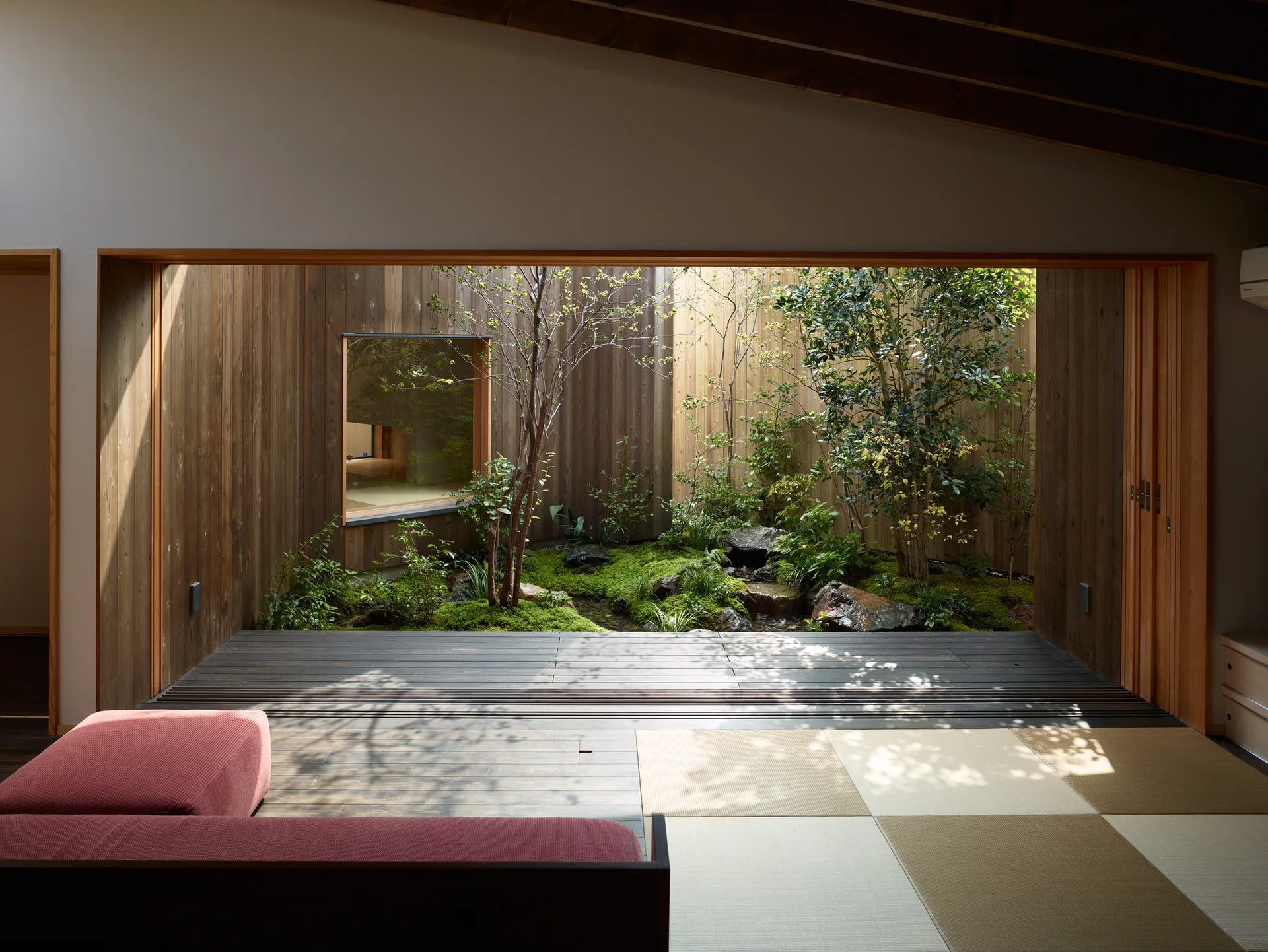 A minimalist living room with tatami mats and a red sofa opens directly onto an enclosed wooden courtyard garden filled with green plants.