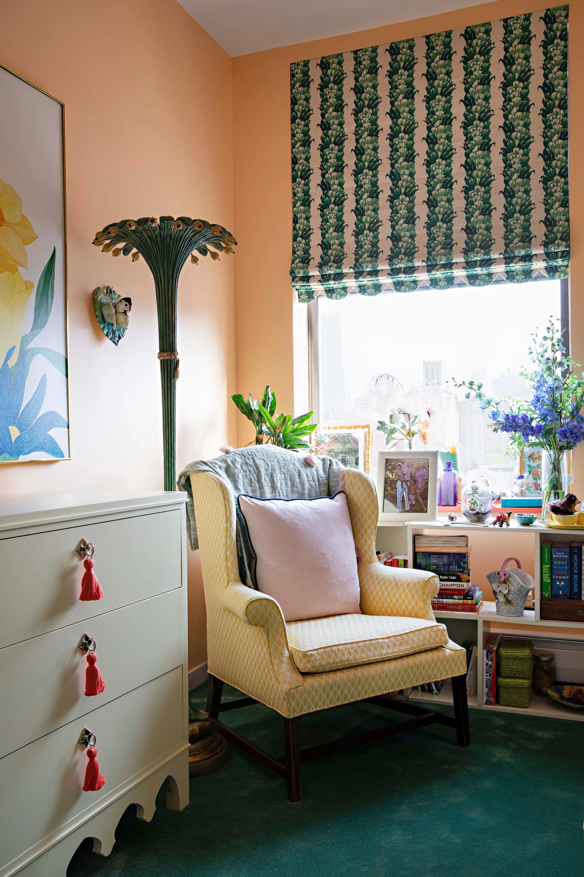 An eclectic reading nook features a yellow patterned wingback chair, peach walls, a cream dresser with coral tassels, and a green botanical roman shade.
