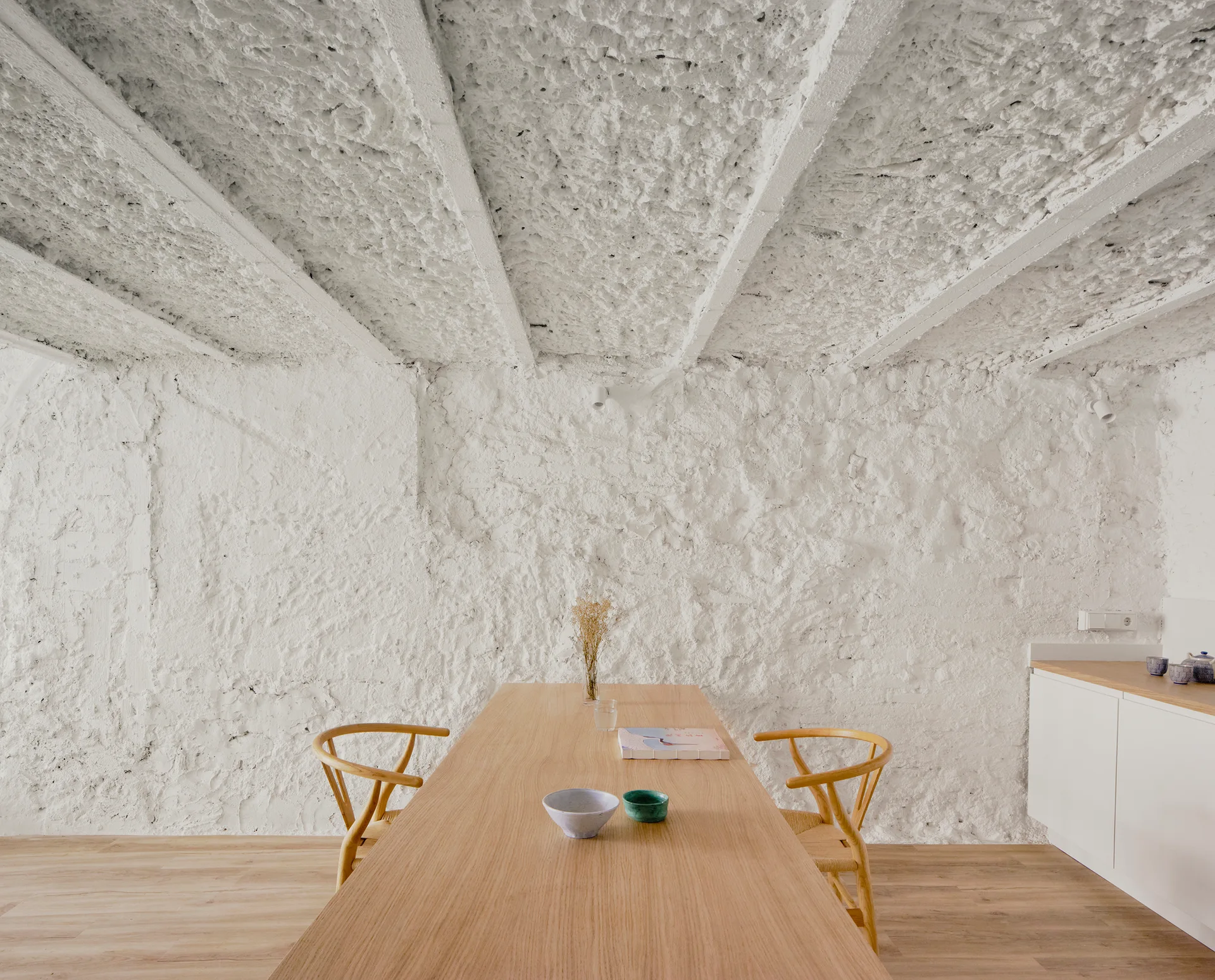 A light wood dining table with two wooden chairs sits in a minimalist room featuring heavily textured white plaster walls and ceiling.