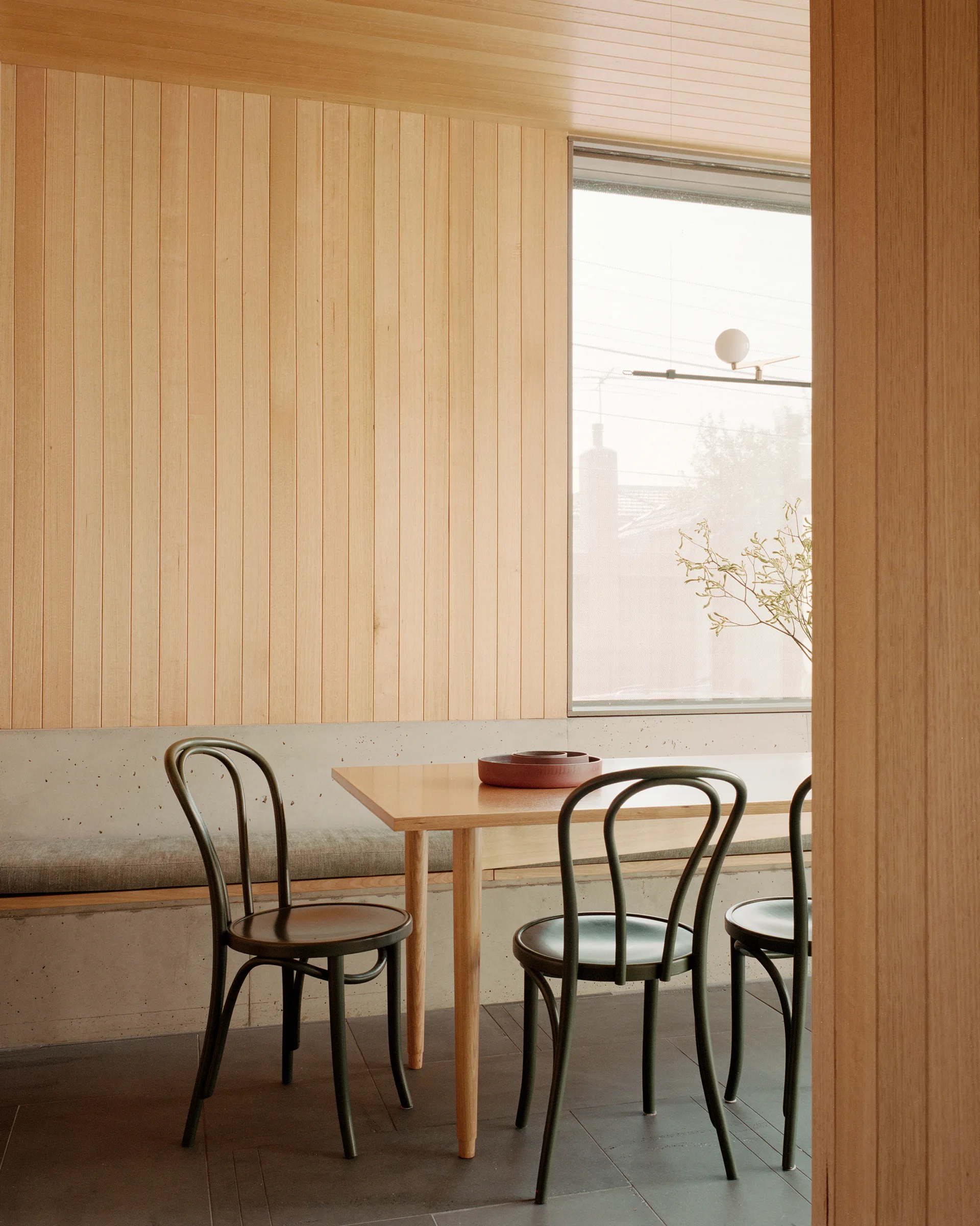 A minimalist dining area features a wood paneled wall, a built-in concrete bench with a cushion, a wooden table, and three dark green bentwood chairs.