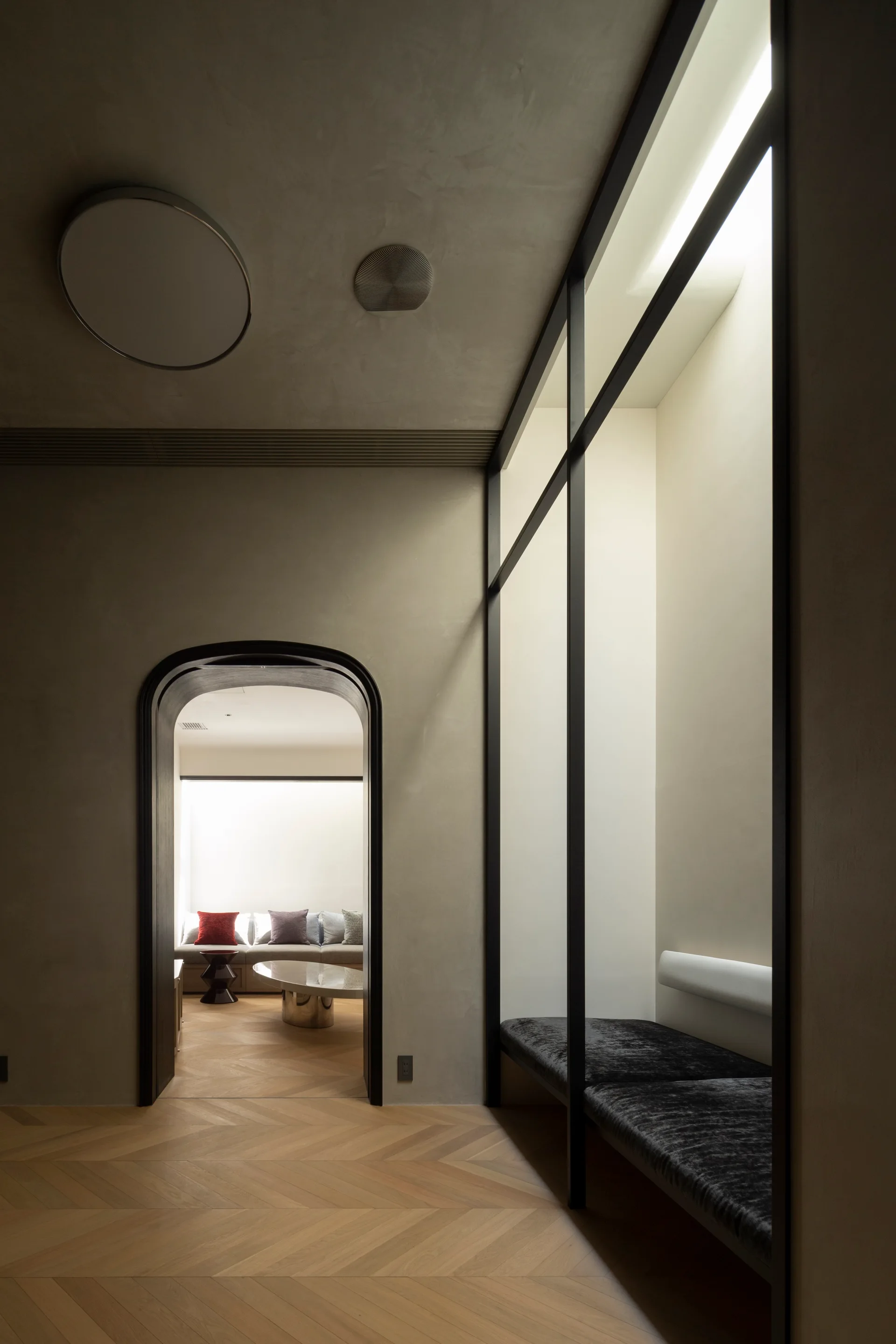 A minimalist hallway featuring a black-framed glass partition with a built-in bench, an arched doorway, and herringbone wood flooring.
