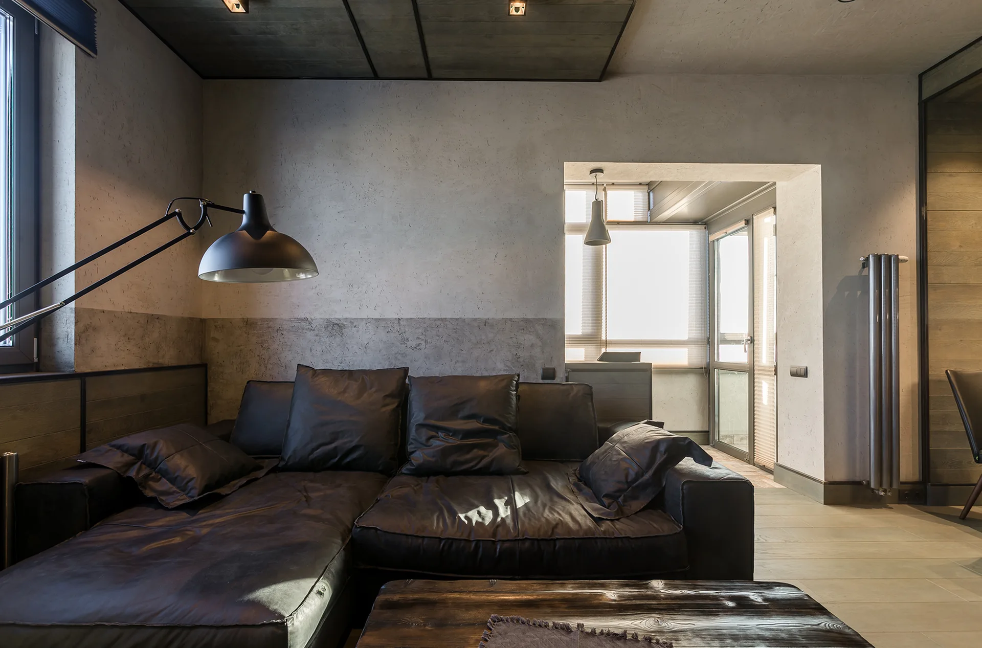 A dark leather sectional sofa sits in an industrial living room with textured grey plaster walls, a black swing-arm floor lamp, and a wooden coffee table.