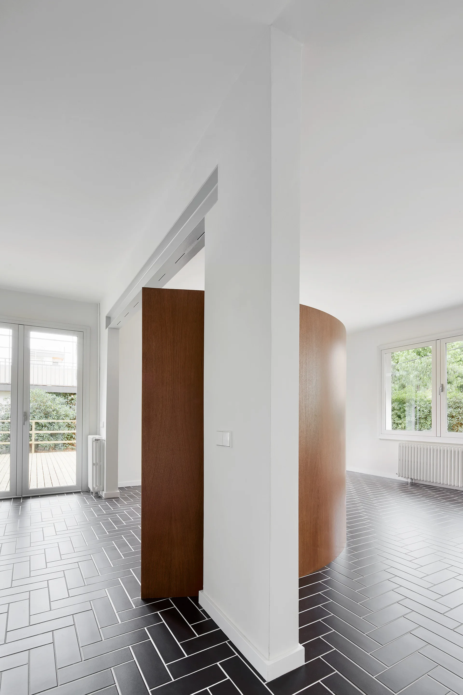 A minimalist room with dark grey herringbone floor tiles, white walls, and a curved warm wood-toned partition wall.