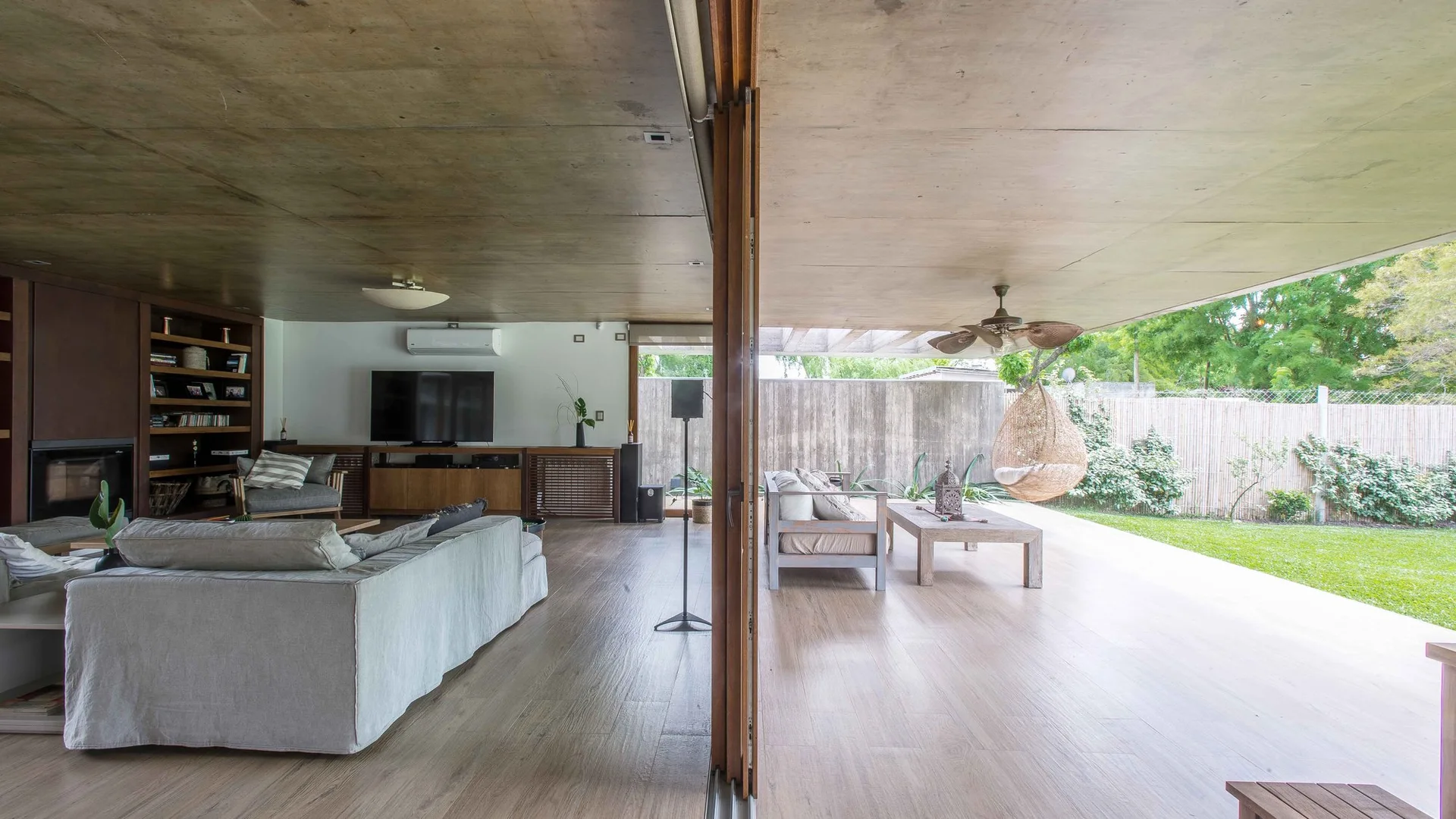 A continuous wood plank floor connects a modern grey living room to an outdoor covered terrace featuring a hanging wicker chair and garden.