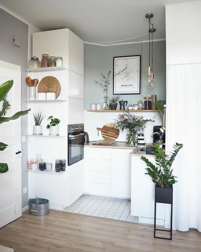 A compact L-shaped kitchen features white cabinetry, a sage green accent wall, light wood countertops, and white floating shelves holding potted plants.