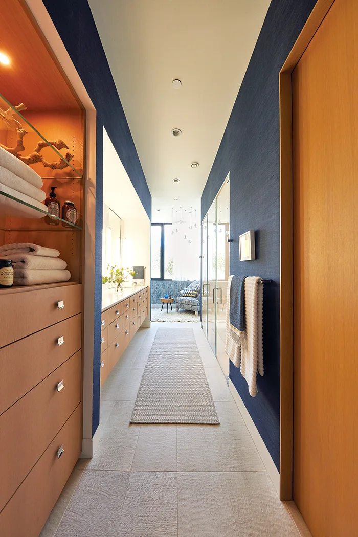 A long bathroom hallway with navy blue grasscloth wallpaper, light tile floors, warm wood cabinetry, and a woven runner rug.