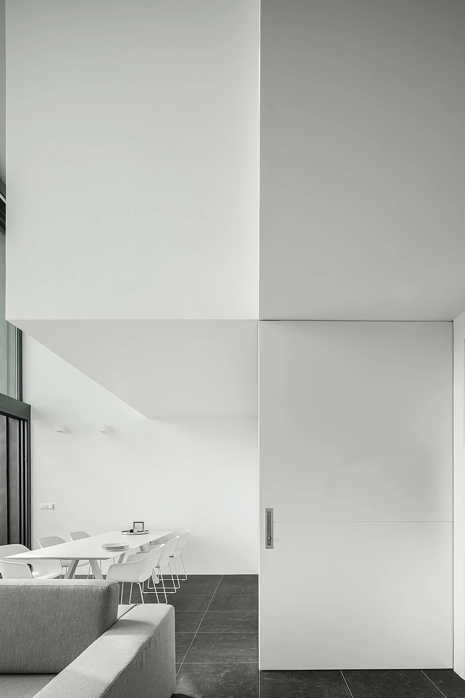A minimalist room with a grey sofa, a white dining set, and large white sliding doors on dark stone floors.