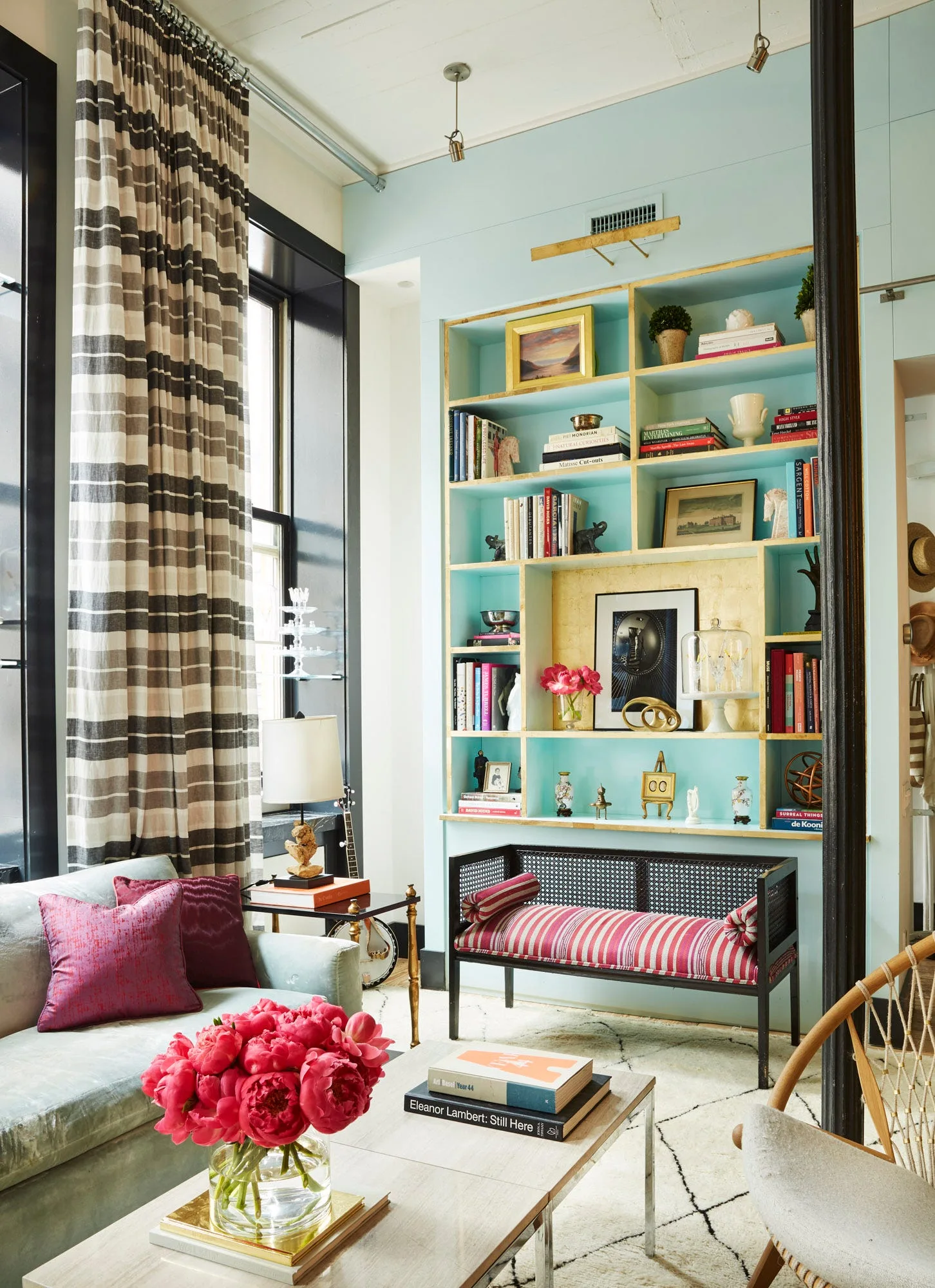 An eclectic living room features a light blue built-in bookcase, a black cane bench with red striped upholstery, and tall black-and-white striped curtains.