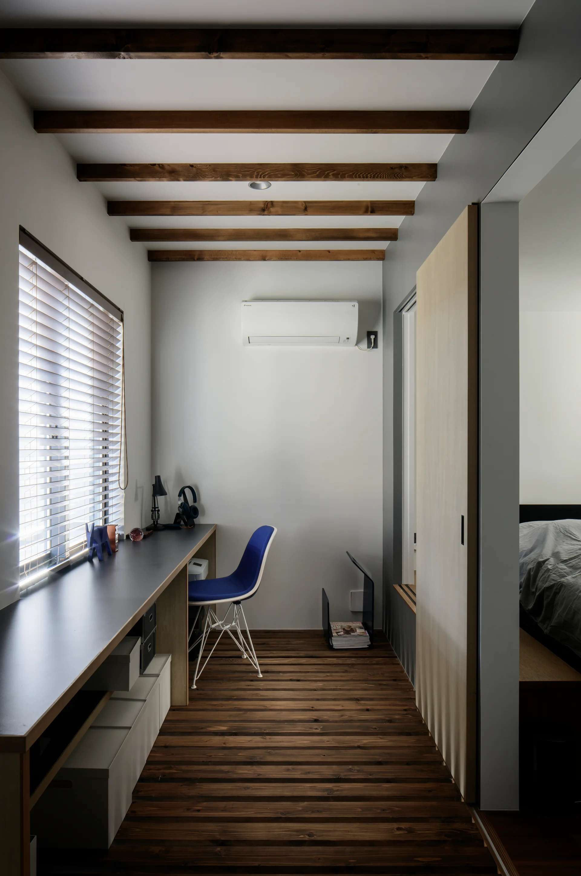 A narrow home office features a long dark desk, a blue chair, exposed wooden ceiling beams, and a dark wood plank floor under a window.