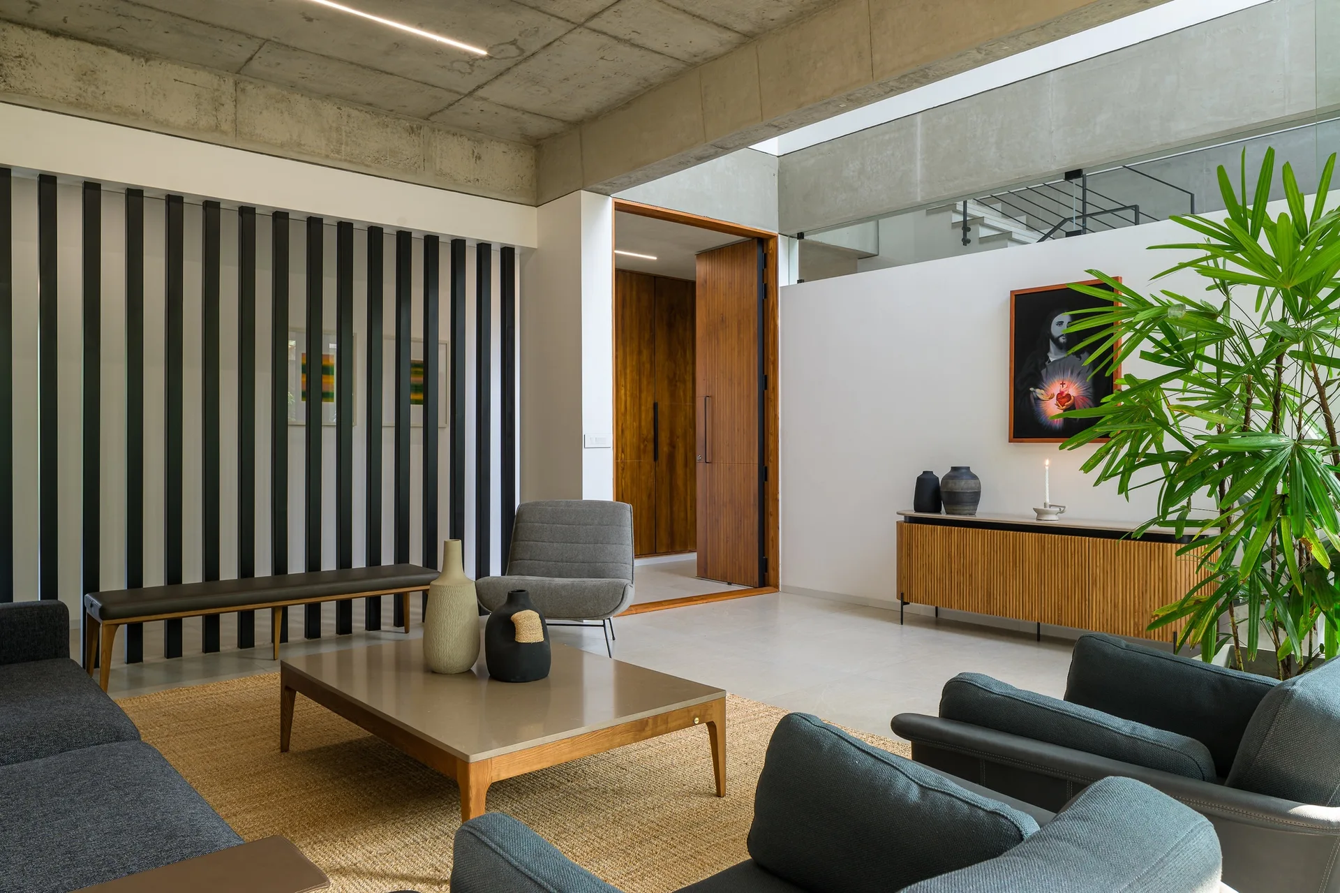 A modern living room features a black-and-white vertical slat wall, an exposed concrete ceiling, grey armchairs, a wooden coffee table, and a potted plant.