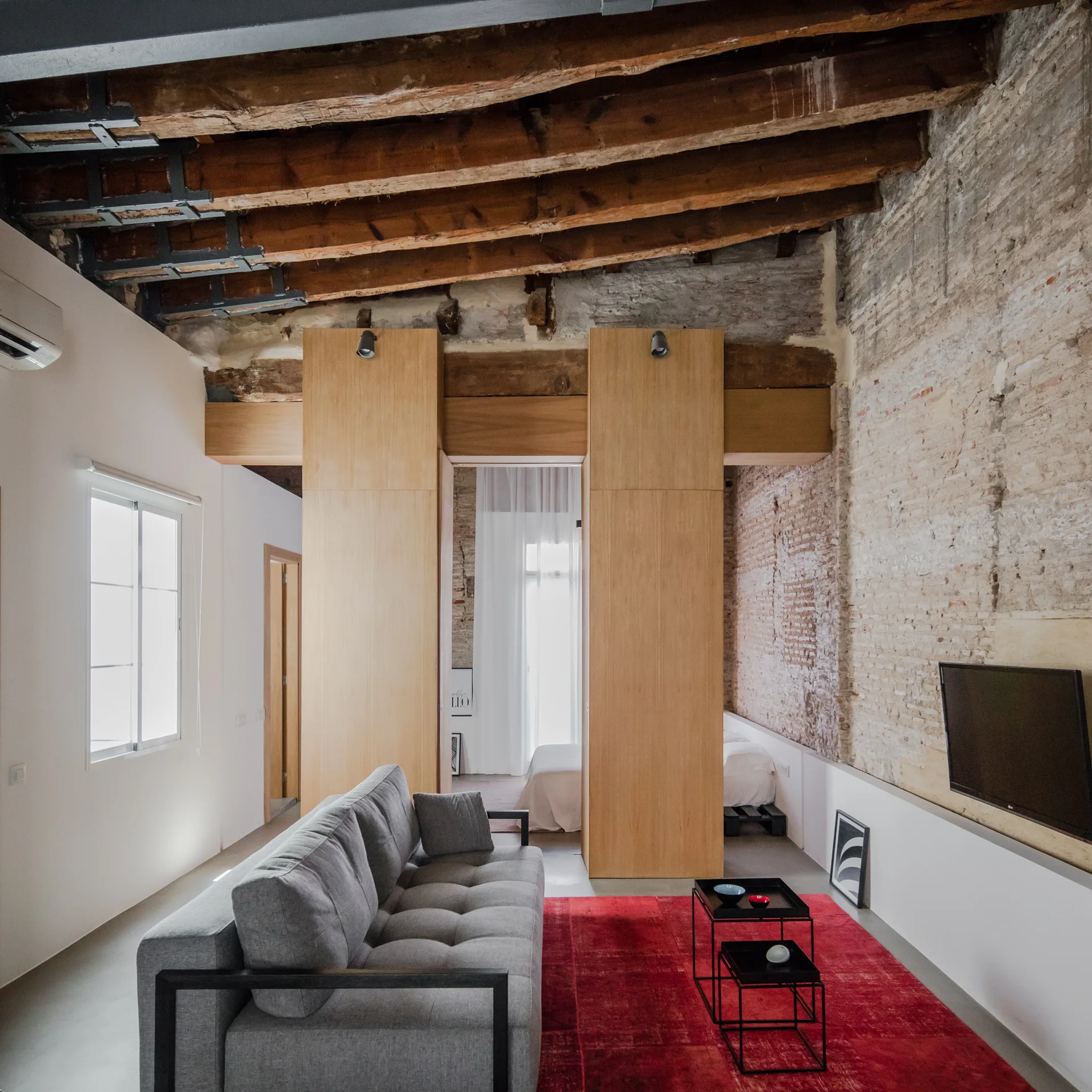 A living room features a grey tufted sofa on a red rug beneath exposed wooden ceiling beams and next to a brick wall.