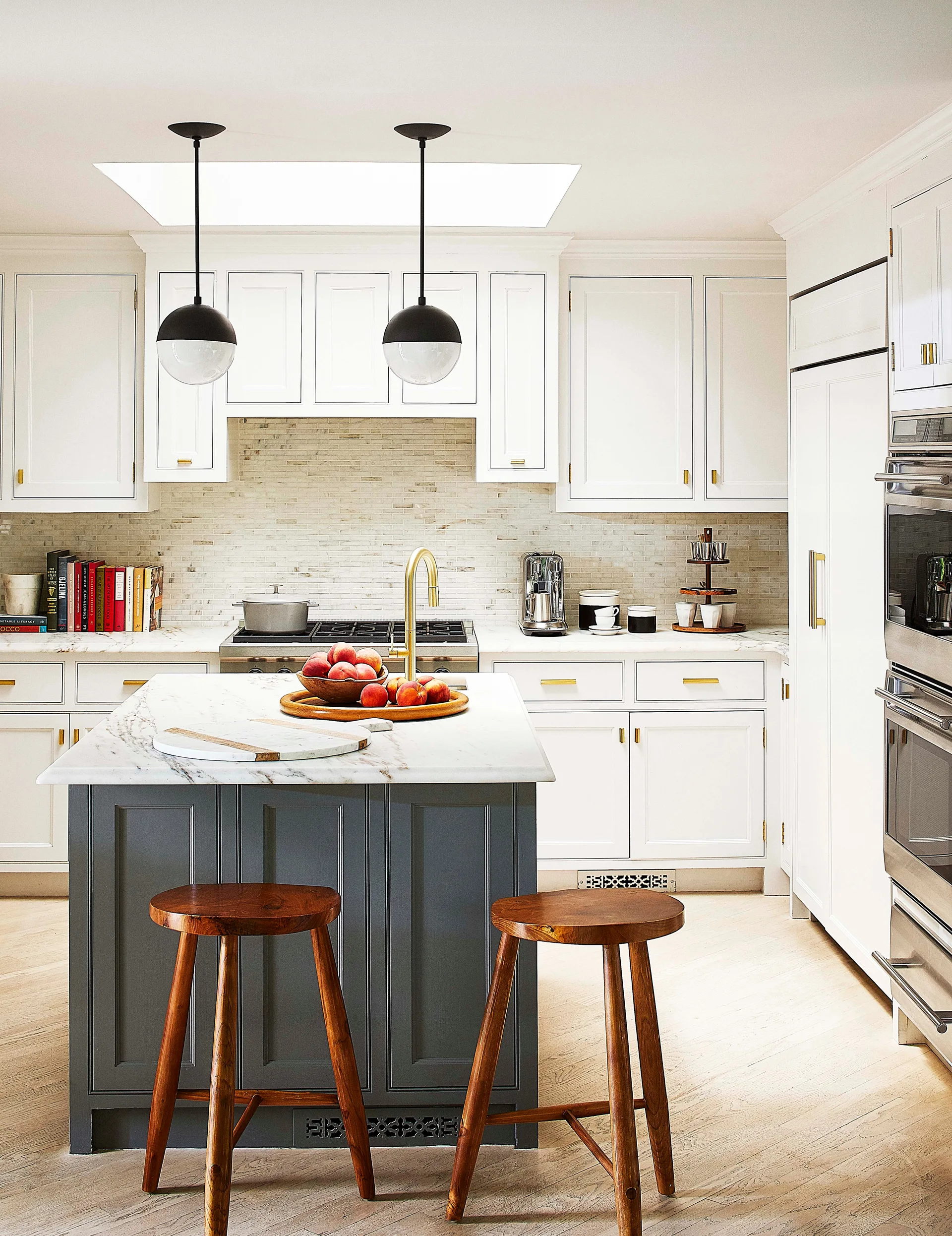 A kitchen featuring a dark gray island with marble countertop, wood stools, white cabinets, and brass hardware under black globe pendant lights.