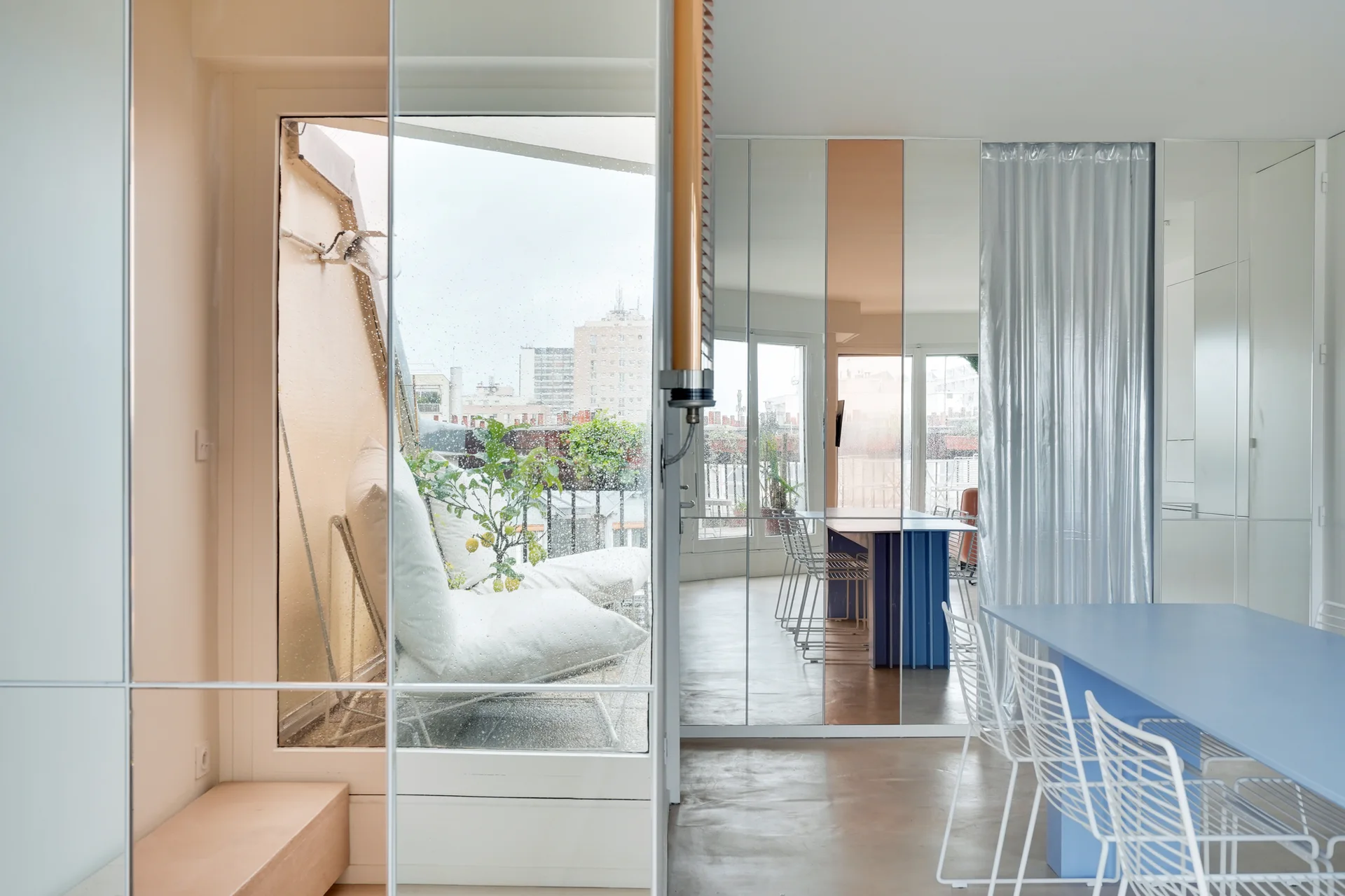 A bright dining room features a blue dining table and white wire chairs next to a large mirrored wall reflecting an outdoor balcony with seating.