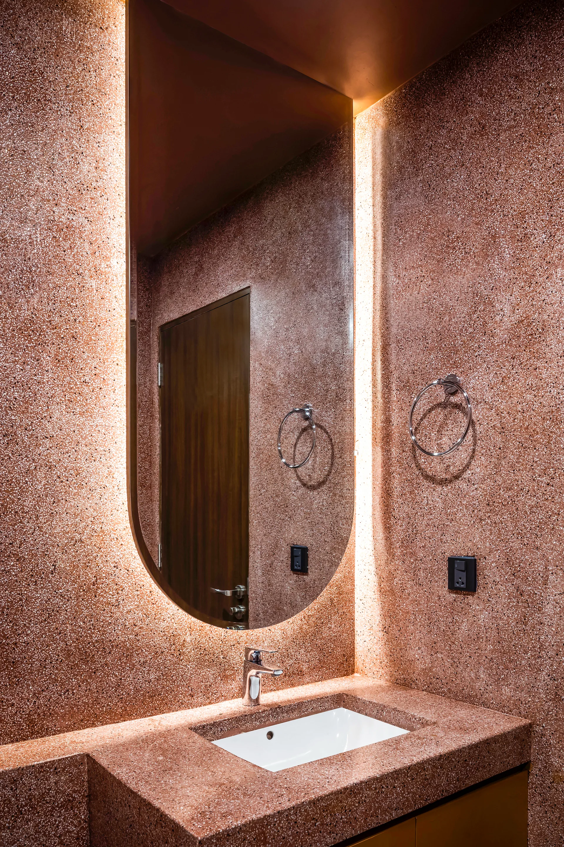 A modern bathroom features a copper-toned, speckled terrazzo vanity with a white integrated sink and an arched backlit mirror.