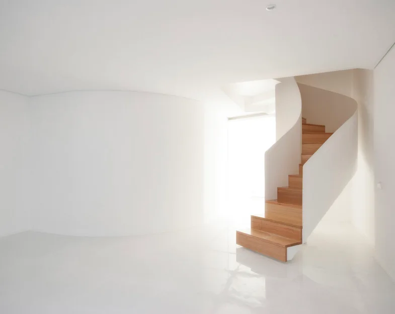 A minimalist white foyer featuring a curved sculptural staircase with natural wood treads and a solid white balustrade on a glossy floor.