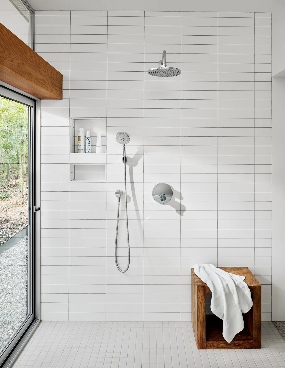 A walk-in shower features stacked white wall tiles, chrome fixtures, a recessed niche, a wooden bench, and a glass door opening to the outdoors.