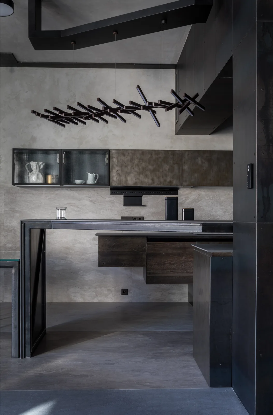 An industrial kitchen features a dark metal island, mesh cabinet doors, textured grey walls, and a black branch-like geometric pendant light overhead.