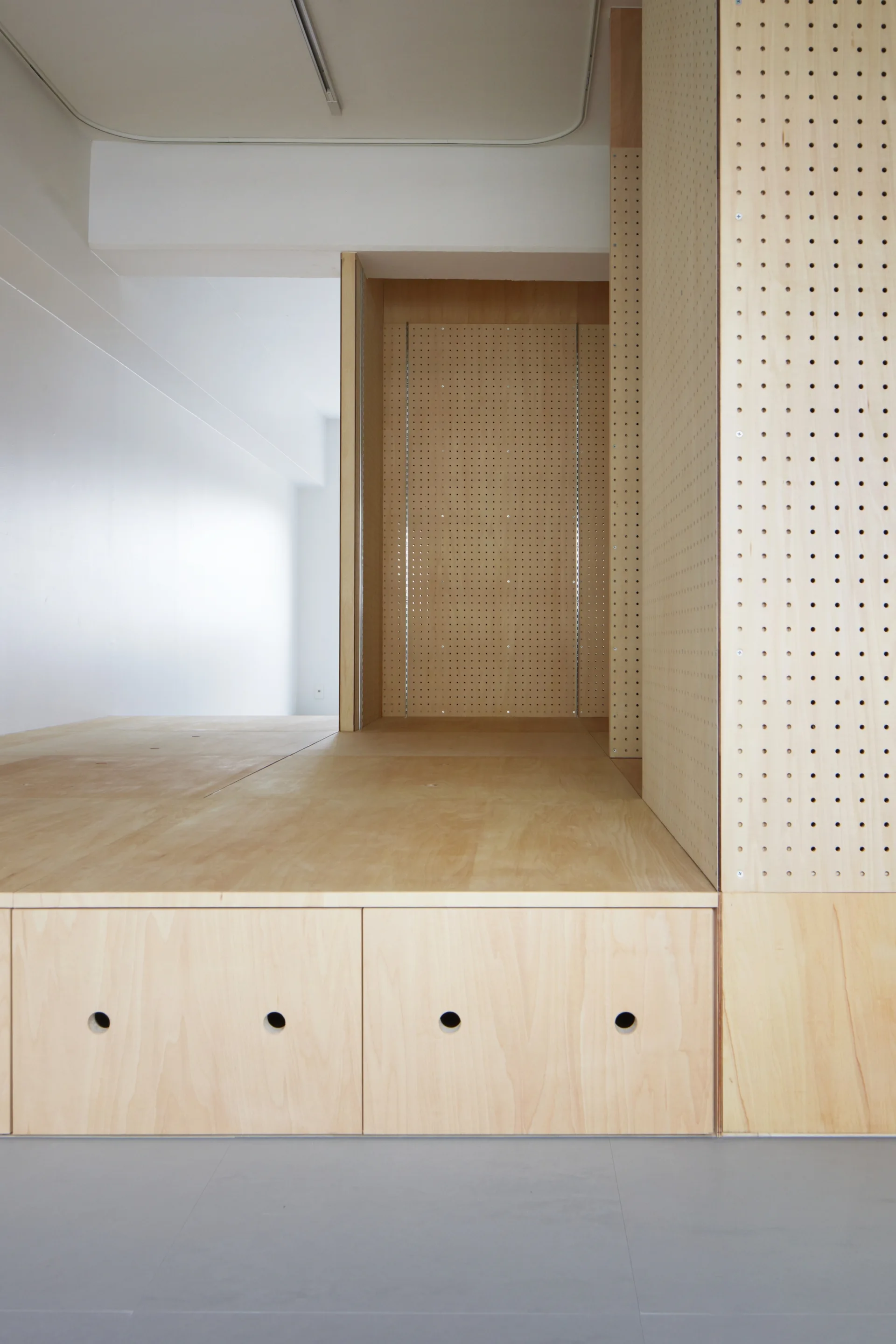 A custom built-in light wood plywood platform features integrated drawers with circular pull holes and vertical perforated pegboard panels.