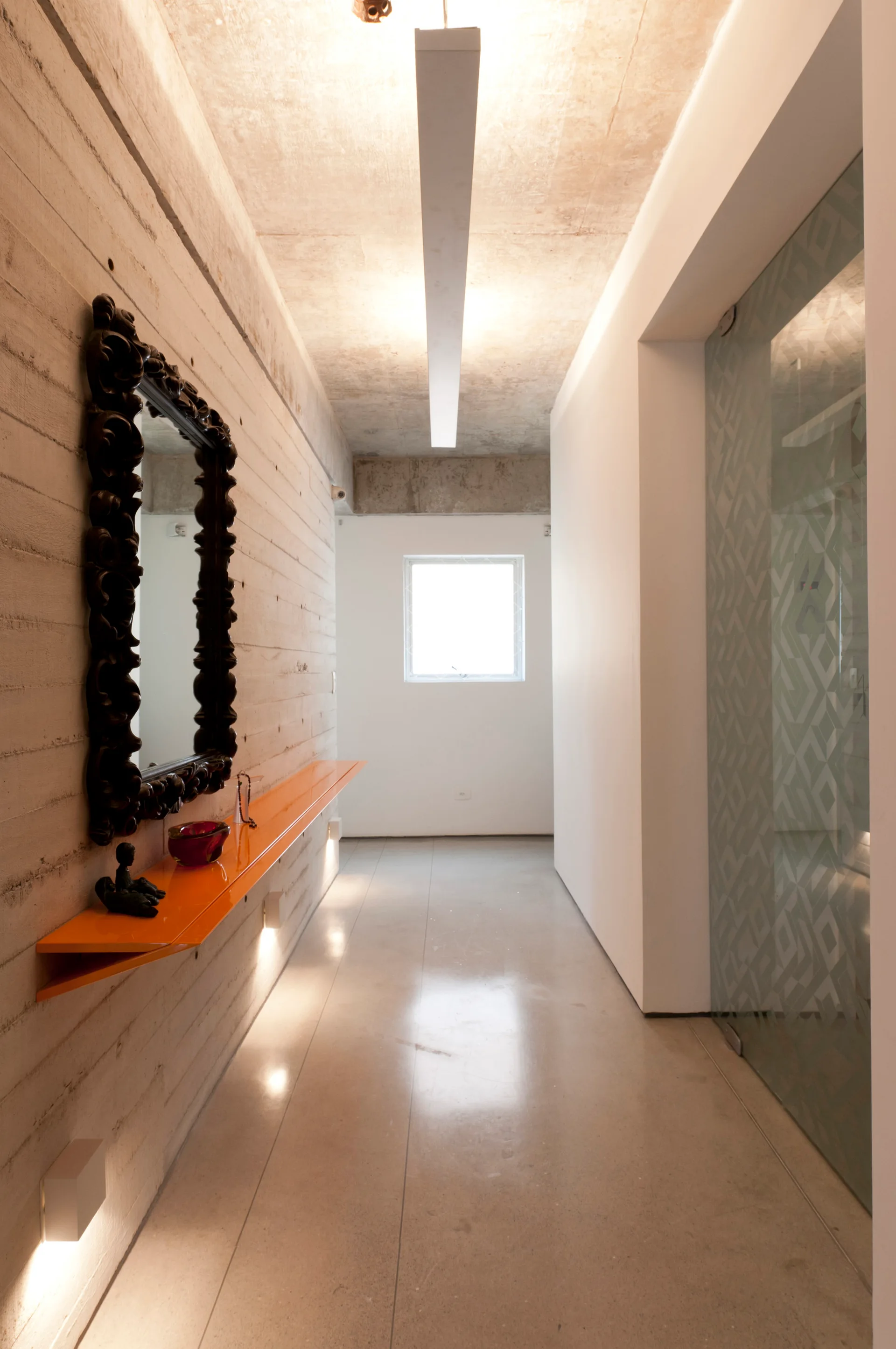 A long concrete hallway features a bright orange floating shelf beneath an ornate black mirror, a polished floor, and a frosted glass door.