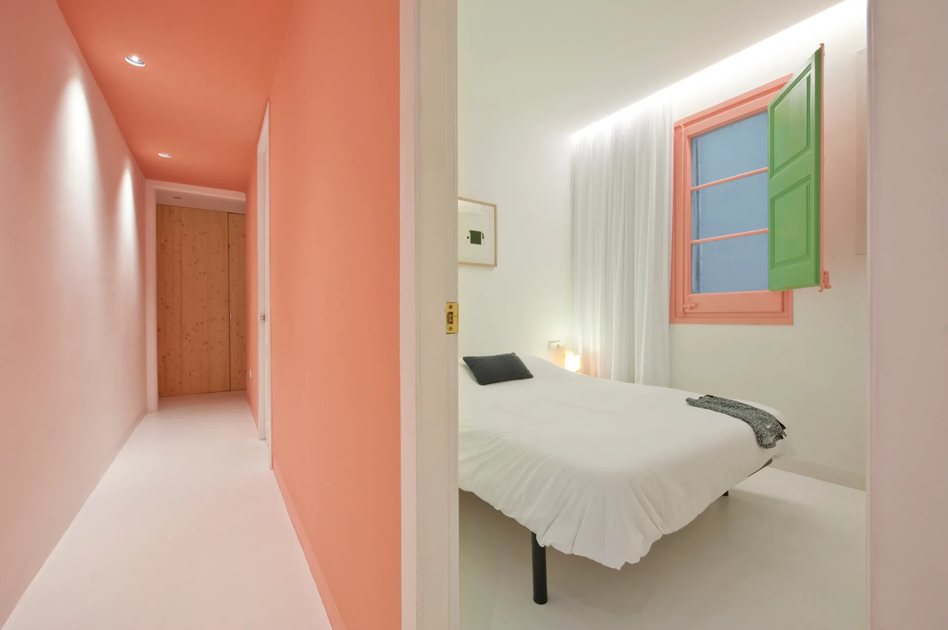 A view looking from a coral pink hallway into a minimalist white bedroom featuring a bed, white curtains, and a window with a green shutter.