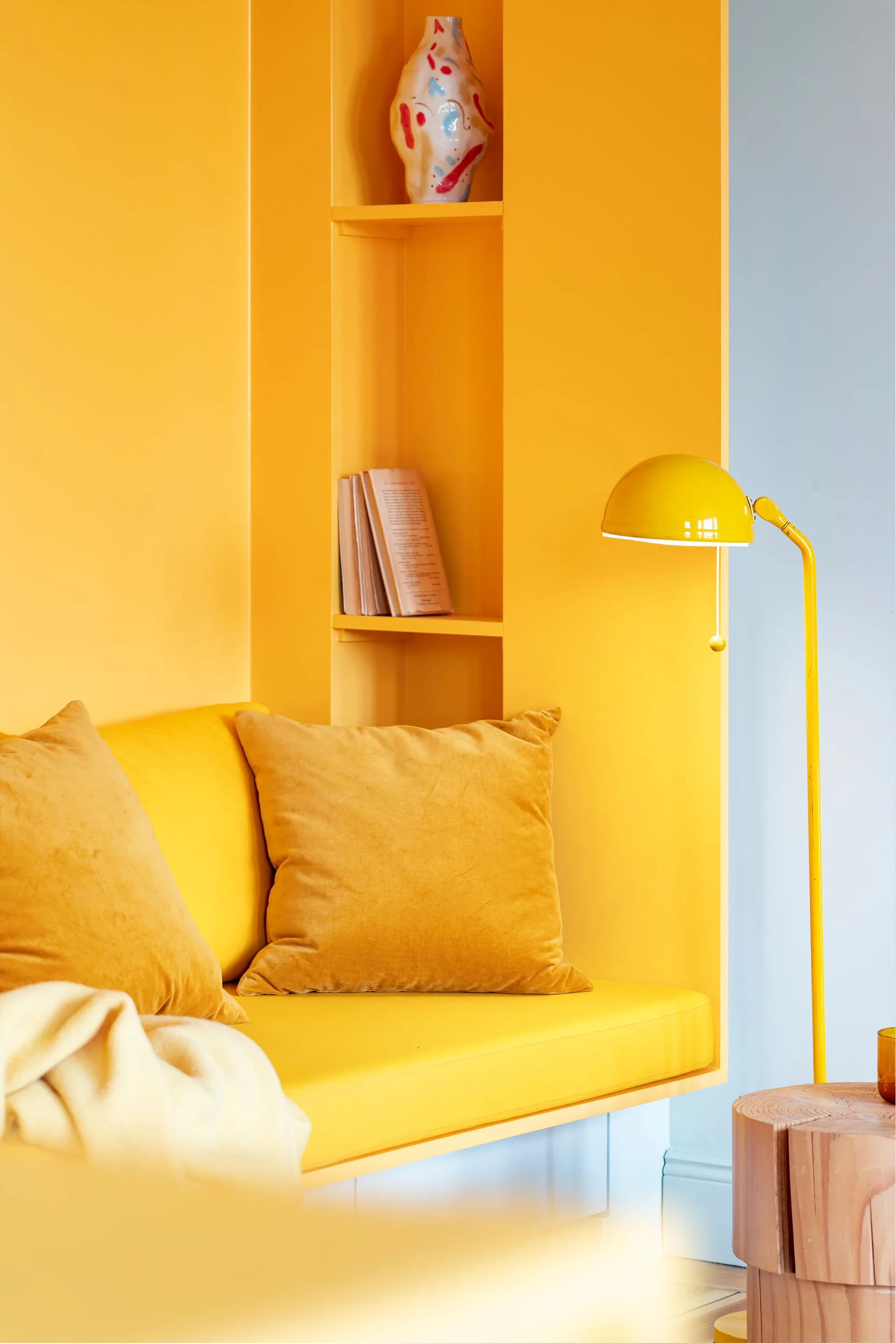 A close-up of a monochromatic yellow built-in seating alcove featuring yellow velvet cushions, a recessed wall shelf with books, and a yellow floor lamp.