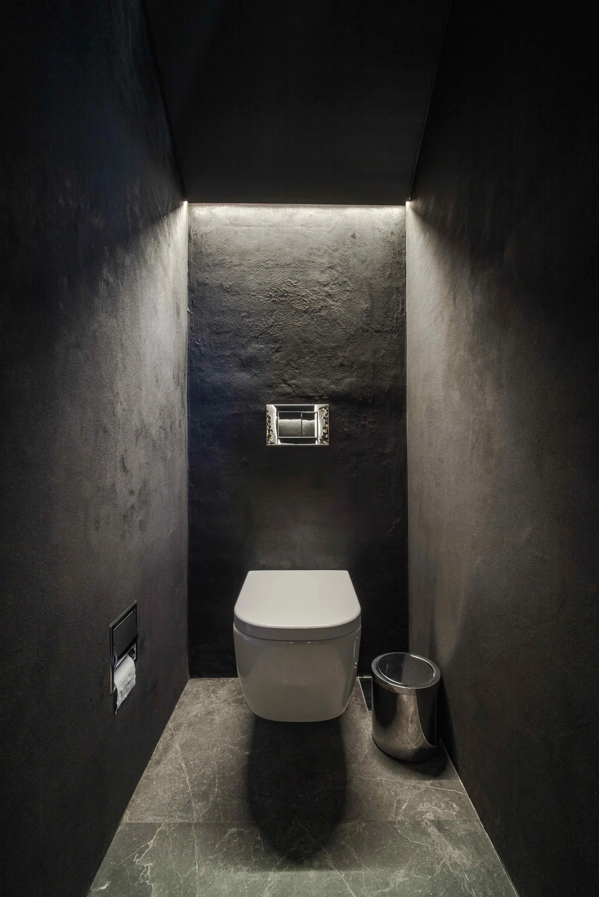 A minimalist black powder room features a white wall-mounted toilet, dark textured walls, and a grey stone floor illuminated by an overhead light strip.