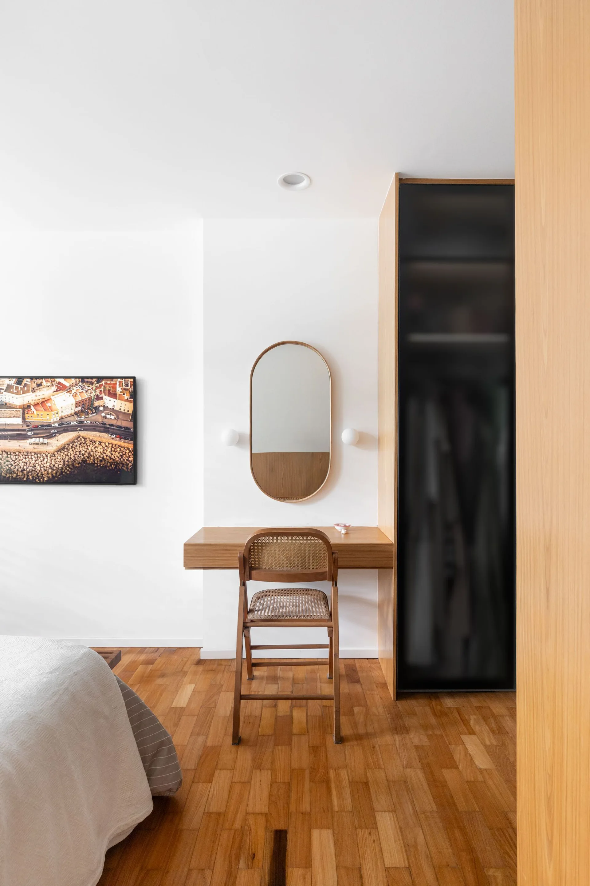 A minimalist bedroom corner features a floating wooden vanity desk, a matching cane-back chair, an oval mirror, and a wood wardrobe with a dark door.