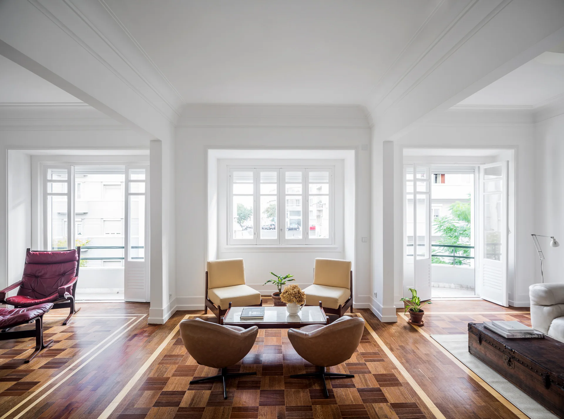 A bright white living room with patterned wood parquet floors, several mid-century modern armchairs, and large shuttered windows.