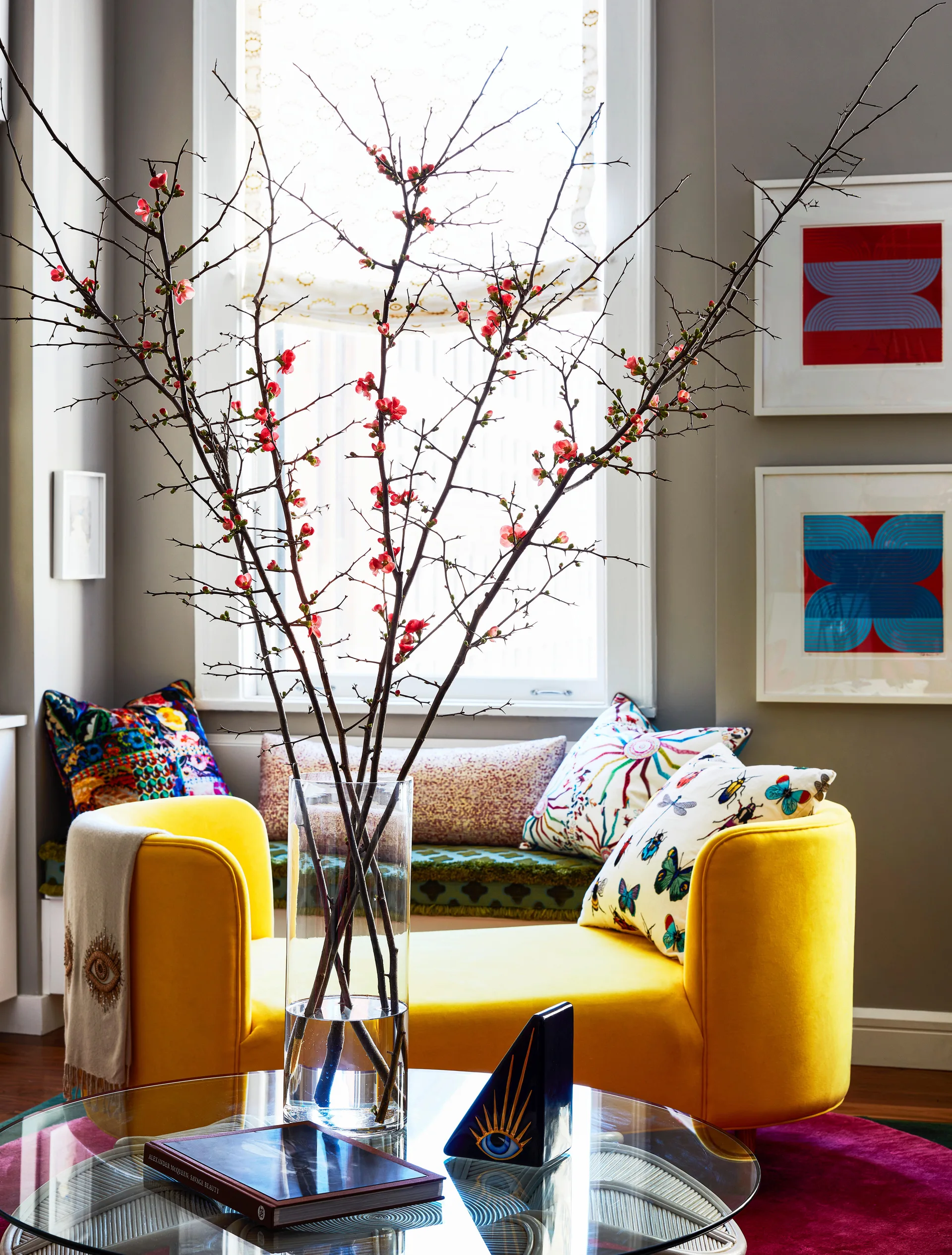 A bright yellow bench accented with colorful patterned pillows sits in front of a window behind a glass table holding tall flowering branches.