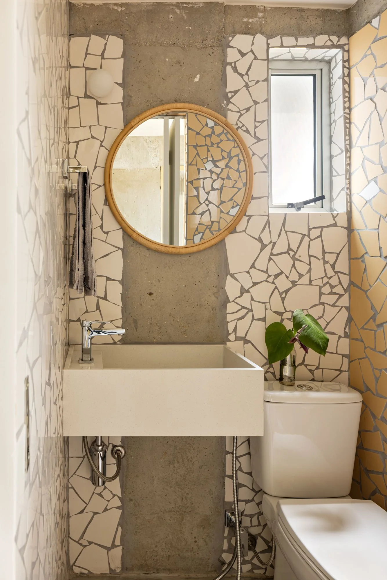 A small powder room features a wall-mounted cream sink, a round wooden mirror, and walls covered in fragmented ceramic mosaic and exposed concrete.