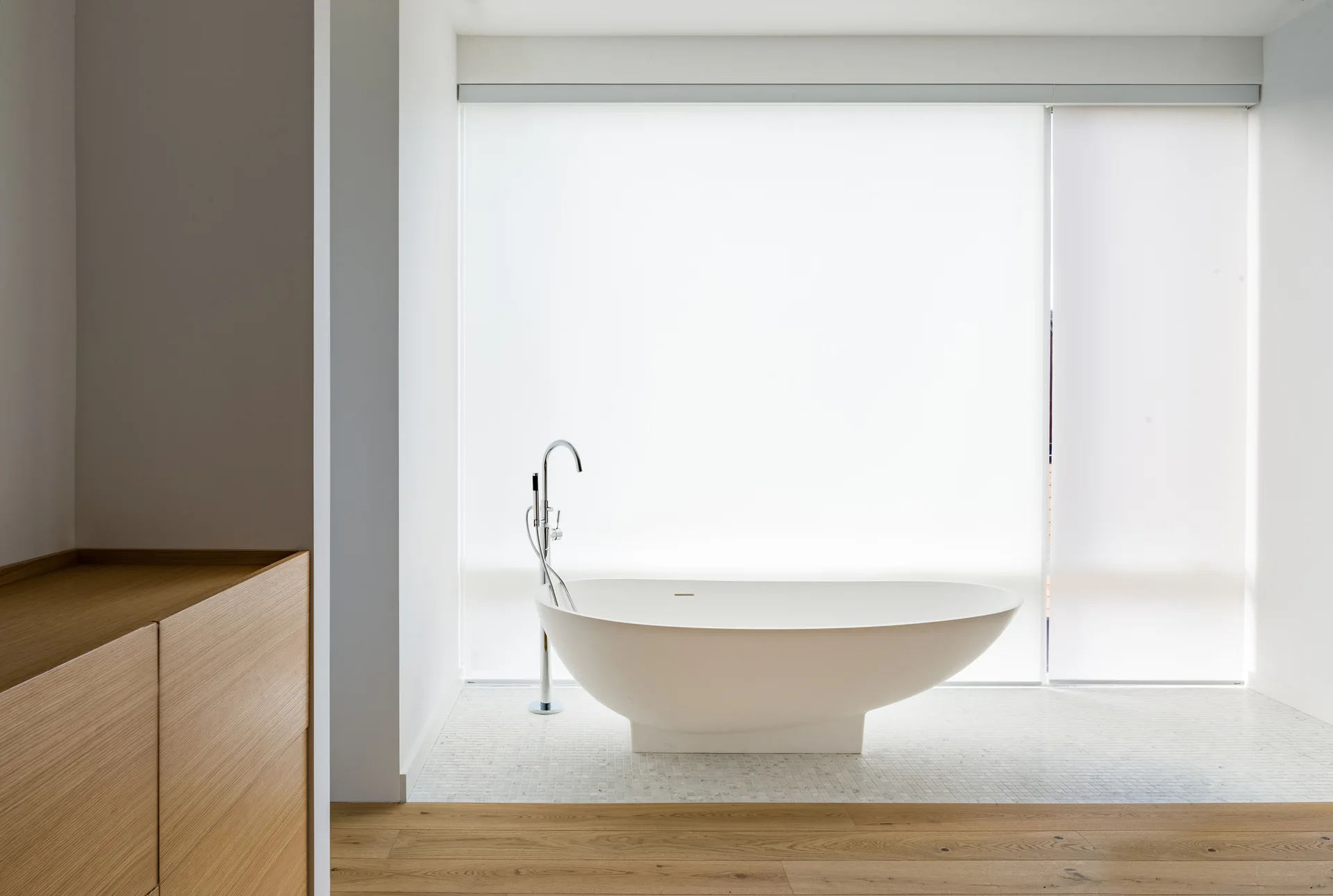 A white freestanding bathtub with a chrome faucet sits on a white mosaic tile floor in front of a large window with roller blinds.