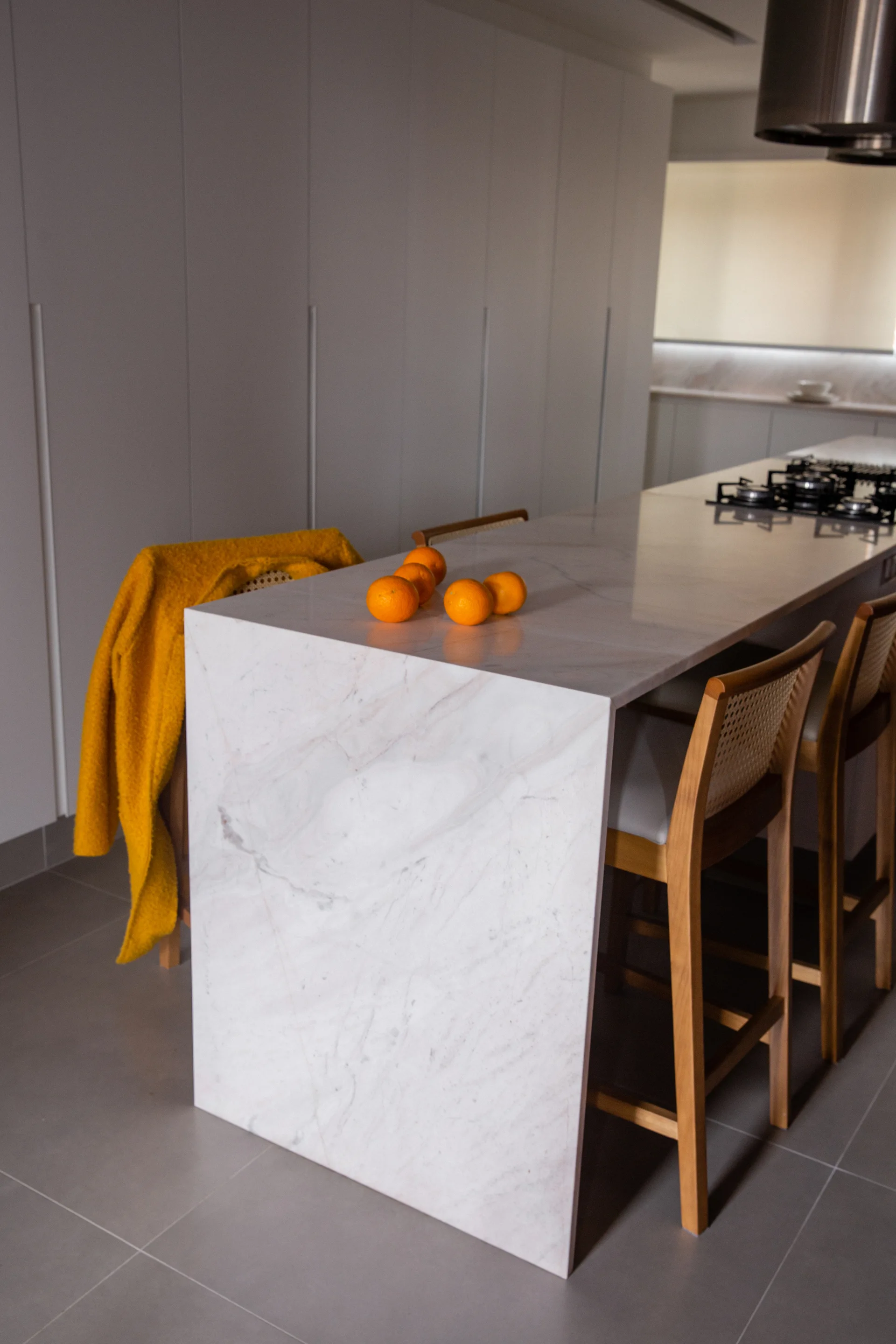 A modern kitchen features a white marble waterfall island with wood bar stools featuring woven cane backs, set against seamless white cabinets.