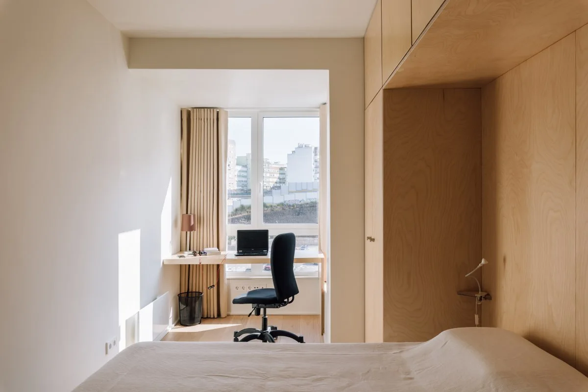 A small minimalist bedroom featuring custom light wood cabinetry, a bed with a beige cover, and a wall-mounted desk inside a window alcove.