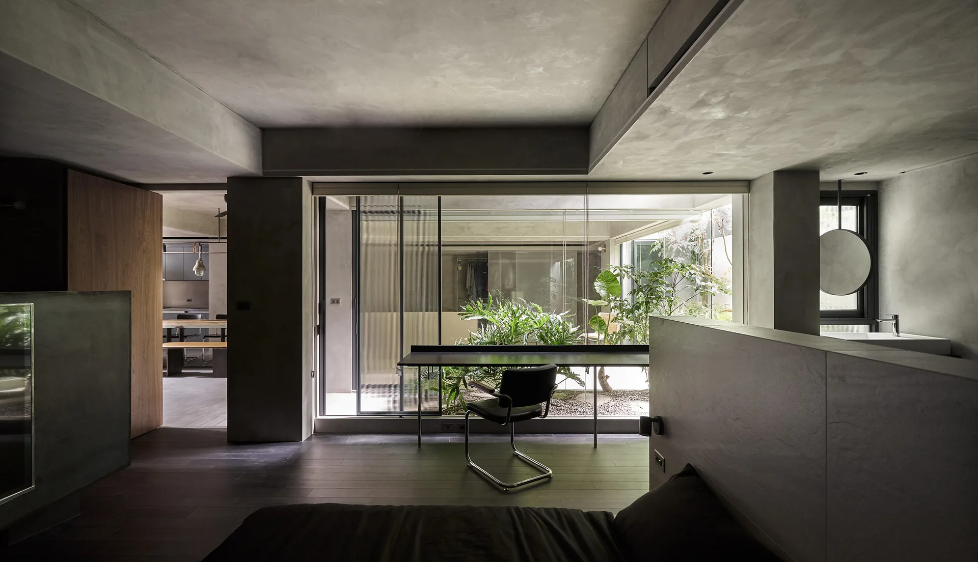 A minimalist bedroom featuring textured gray plaster walls, a slim black desk, and a large window looking out to a green courtyard garden.