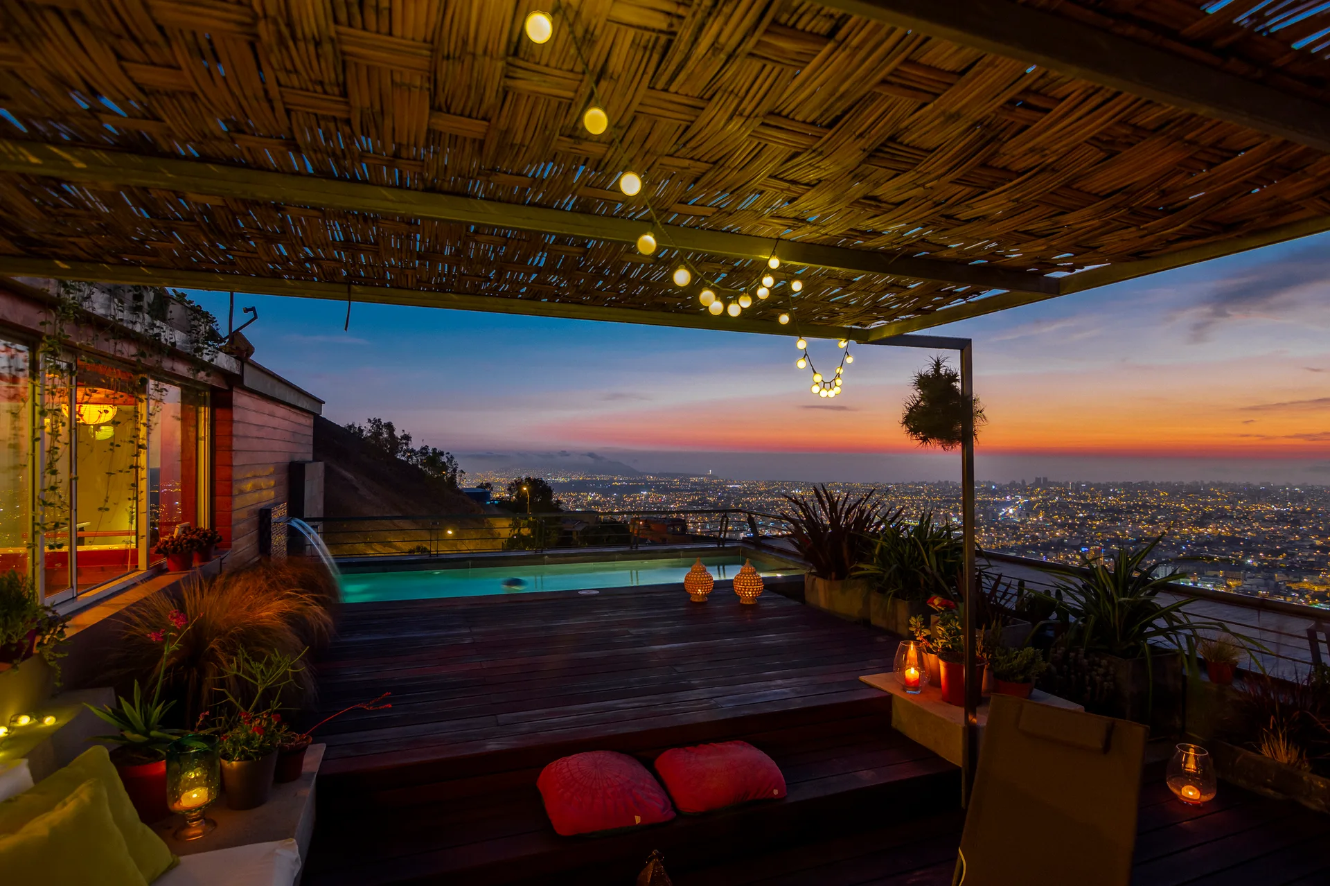 A wooden rooftop terrace featuring a swimming pool, woven bamboo overhead canopy with string lights, and a view of the city skyline at sunset.