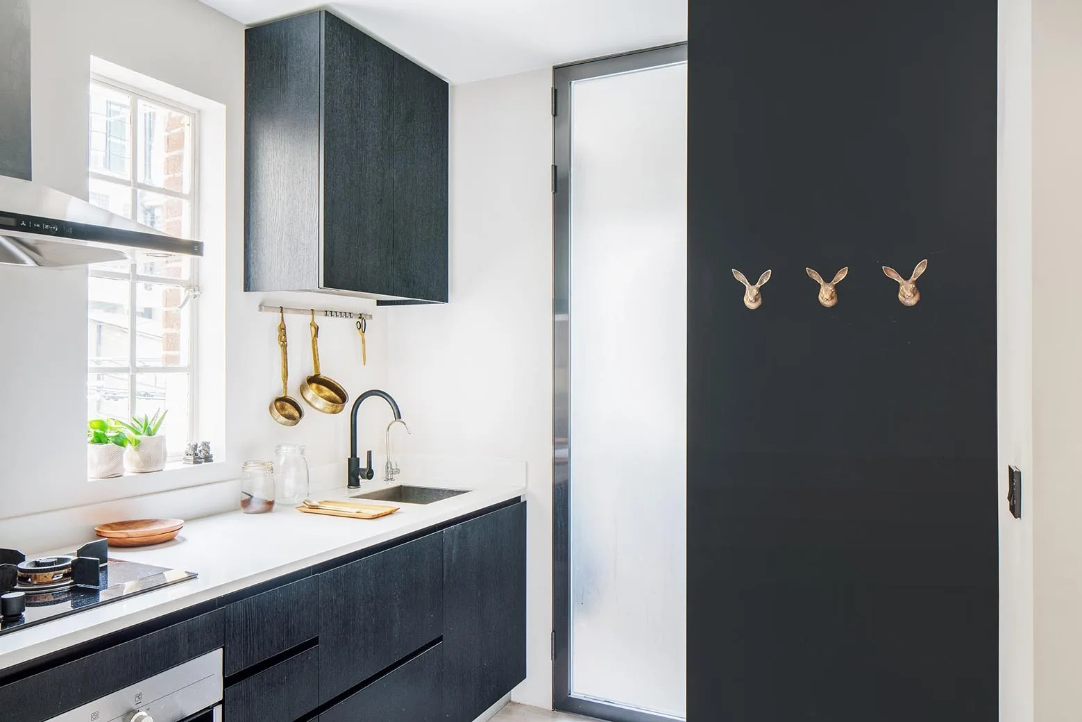 A modern kitchen with matte black cabinetry, white countertops, and three brass rabbit hooks mounted on a black accent wall.