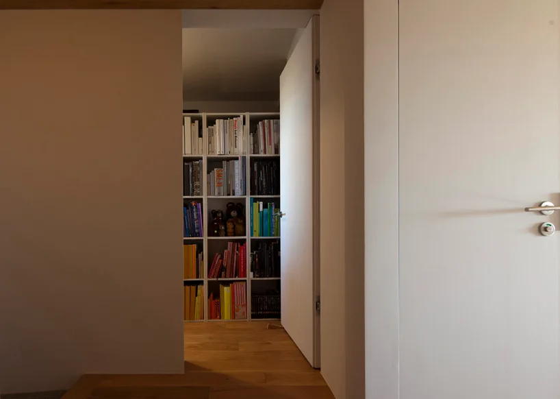 A view through an open minimalist white door reveals a tall grid bookshelf filled with colorful books on a light wood floor.